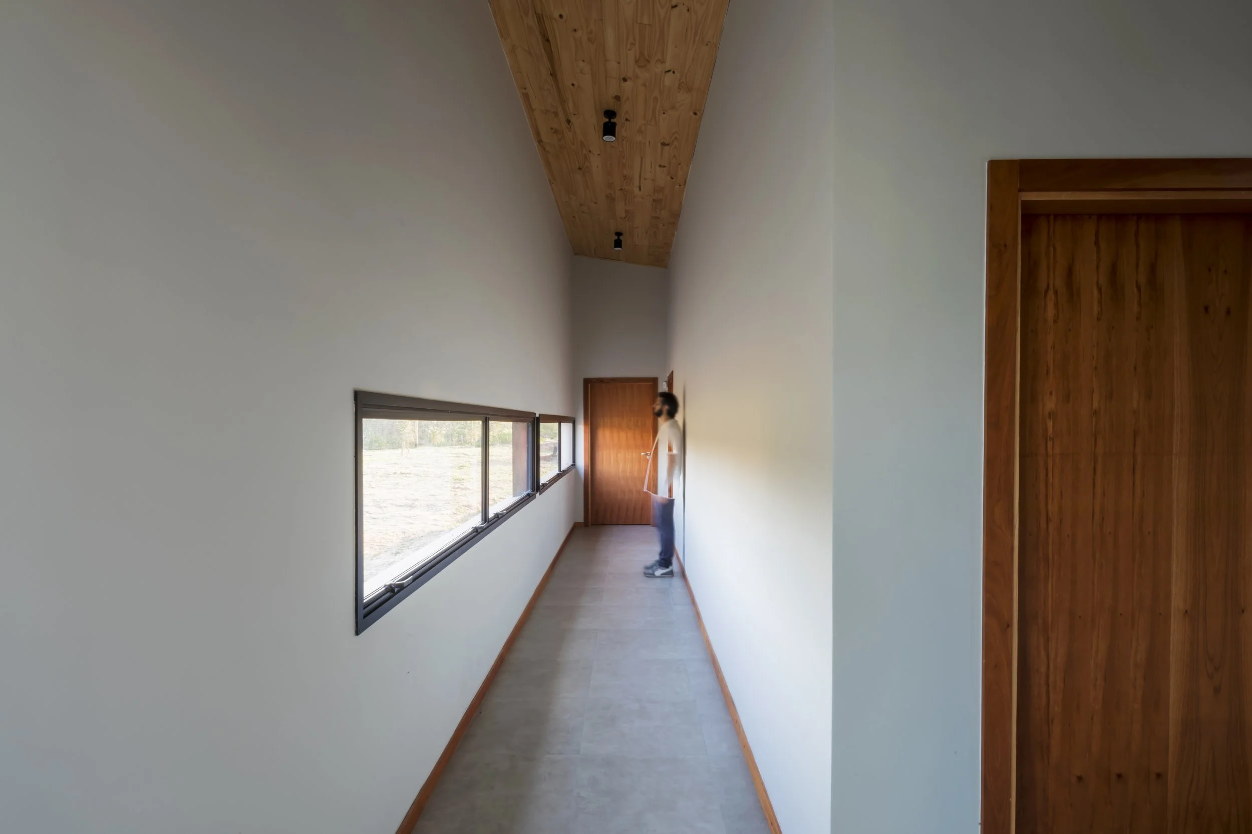 PRO ARQUITECTURA PARAGUAY architecture design PROSA 
Pasillo interior de una casa con ventana larga y techo de madera, una persona de pie mirando hacia la puerta, paredes blancas y suelo de madera.