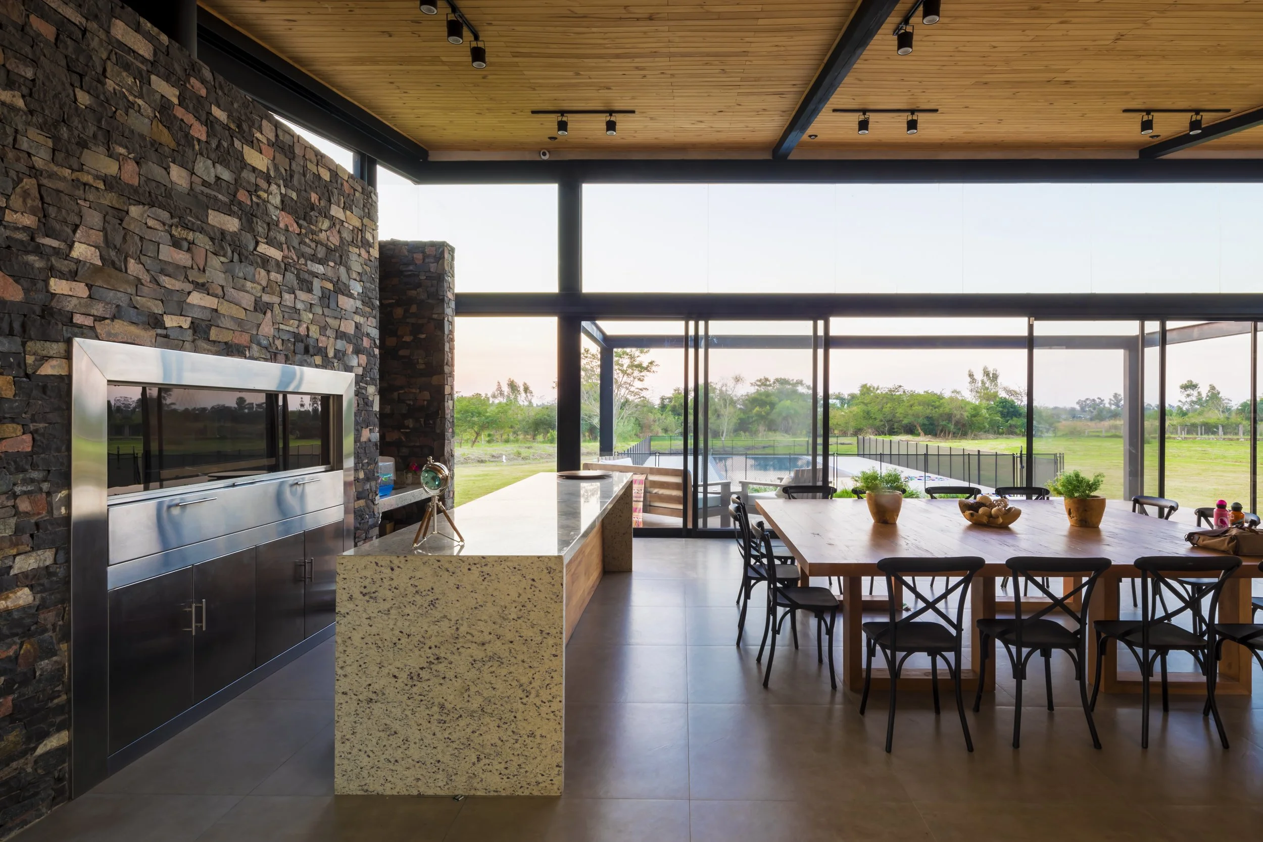 Interior de una casa moderna con vista al jardín, comedor grande con mesa de madera y sillas negras, cocina con encimera de granito y pared de piedra, grandes ventanas de vidrio que dejan ver un campo verde.