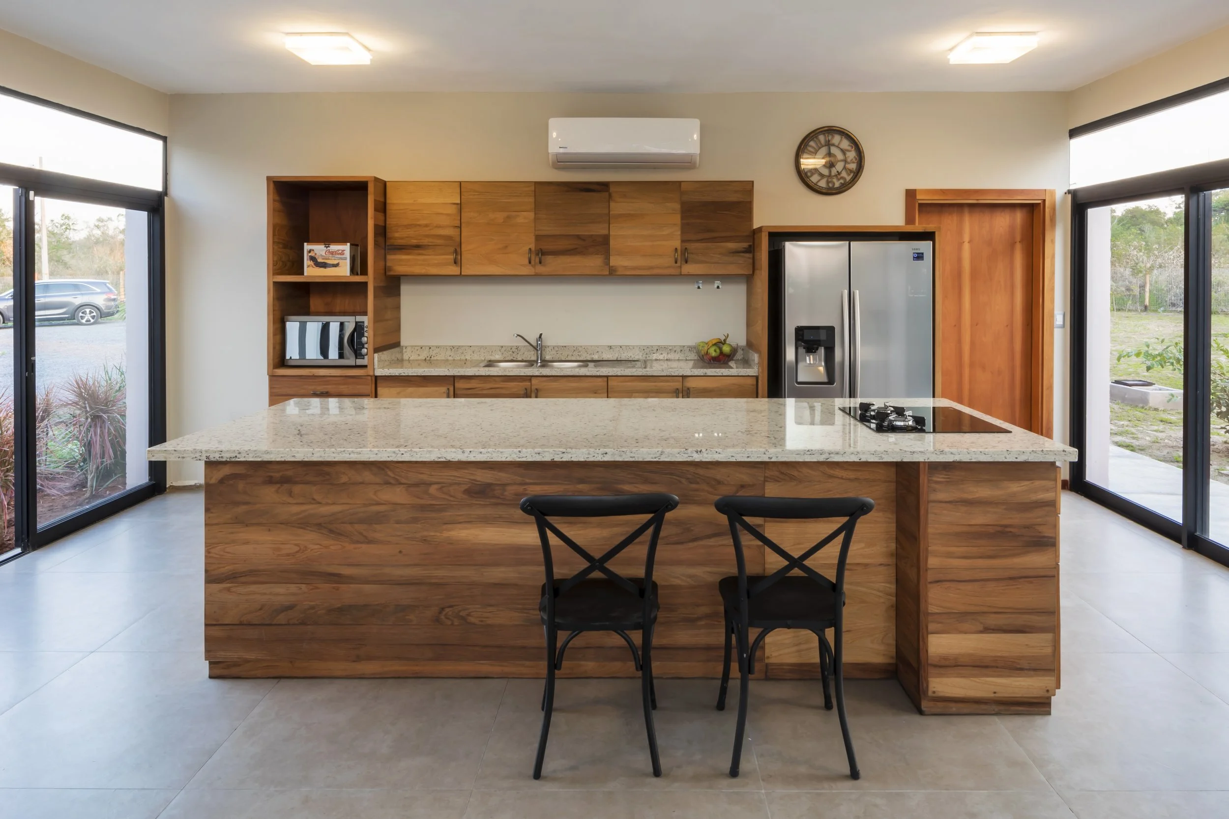 Cocina moderna con encimera de granito, electrodomésticos de acero inoxidable y armarios de madera, con ventanas grandes que dejan entrar luz natural.