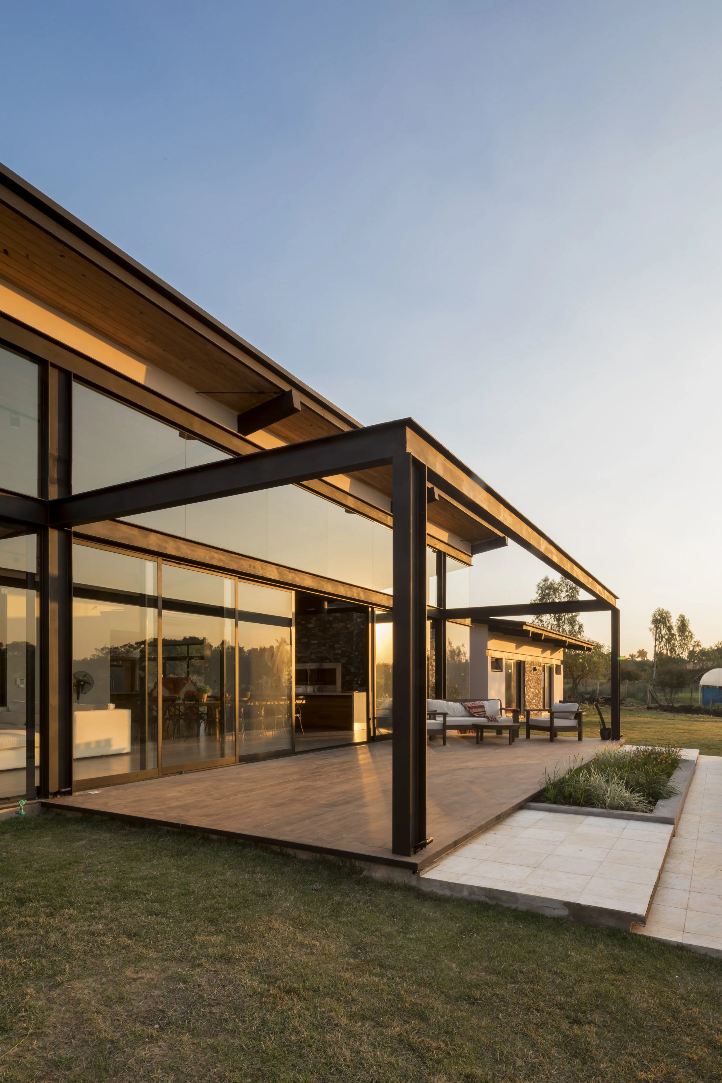 Vista de una casa moderna con grandes ventanales y patio al atardecer, con muebles de exterior y jardín.