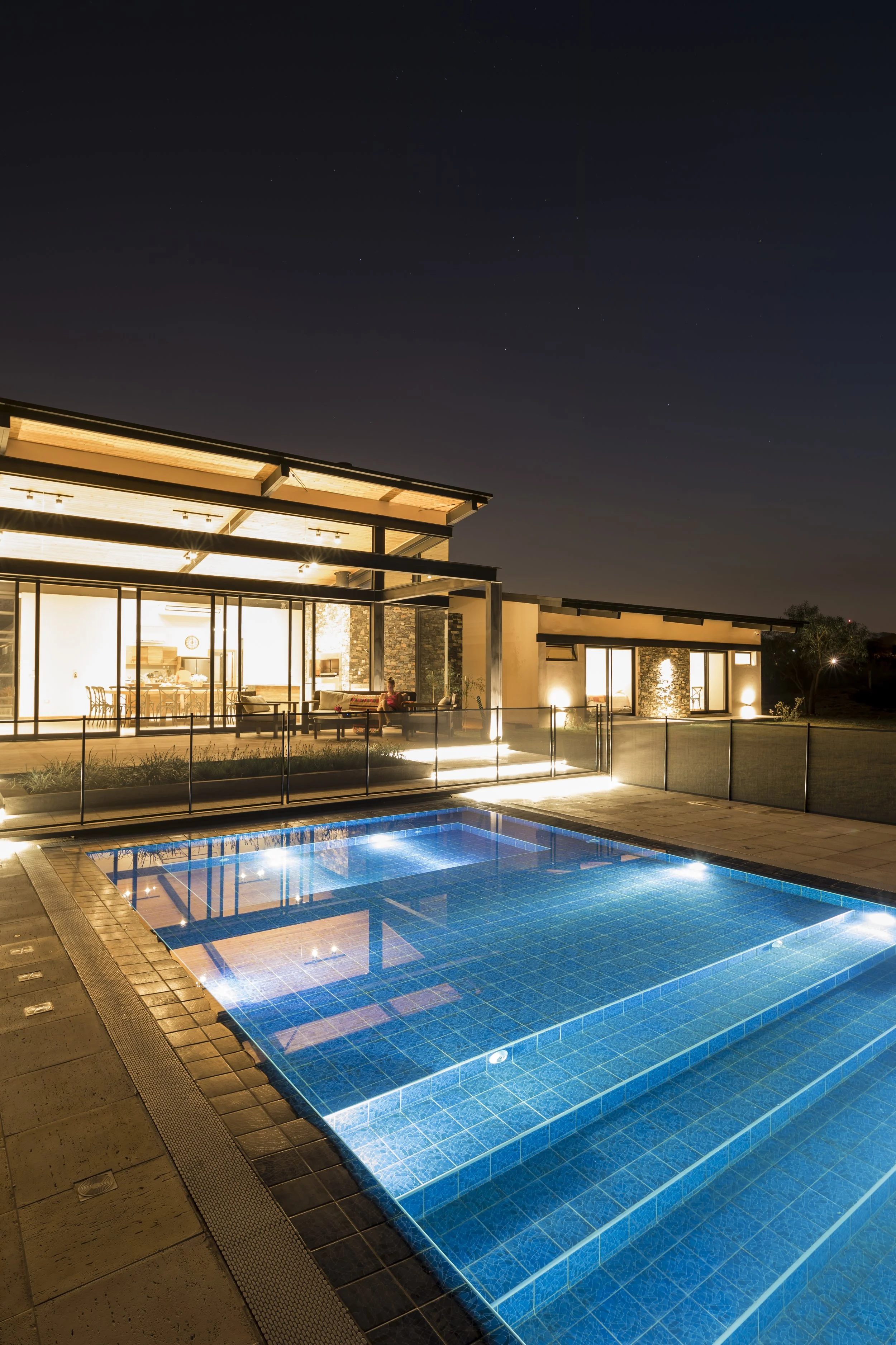 Casa moderna con iluminación, piscina con luces, patio y áreas exteriores en la noche.