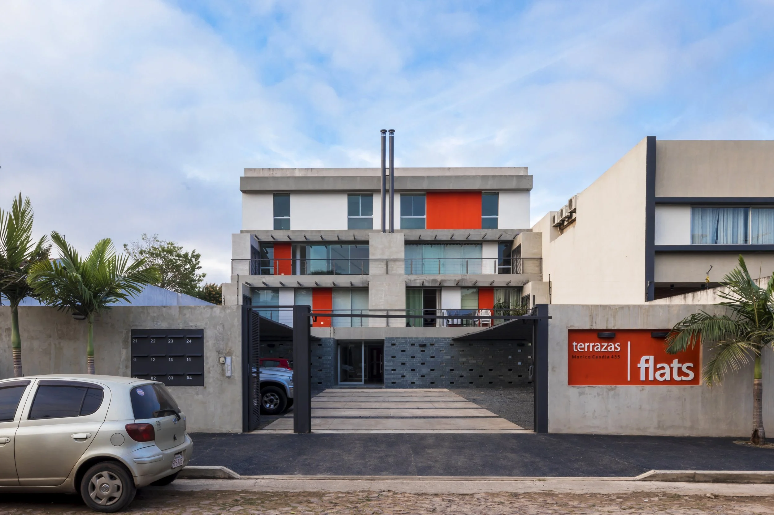 Edificio moderno de apartamentos con fachada de concreto, balcones de vidrio y señal de alquiler de flats en la calle, con carros estacionados y palmeras a los lados.