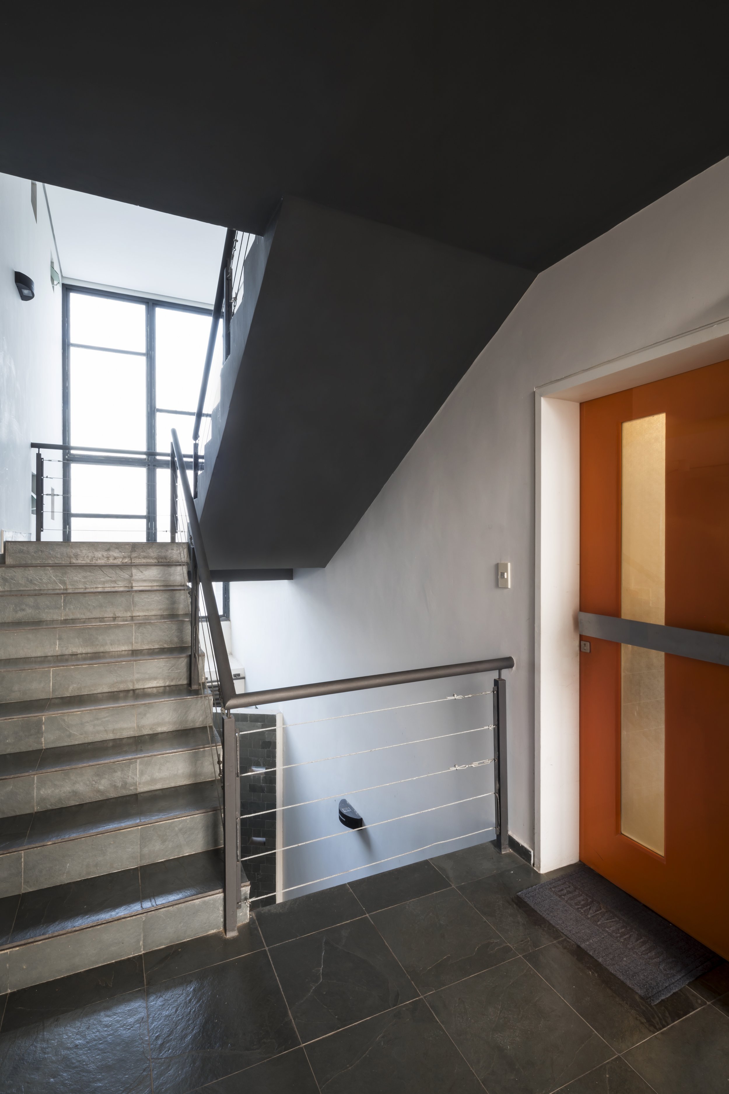Escalera y pasillo en un edificio moderno, con puerta de color naranja y barandilla metálica.