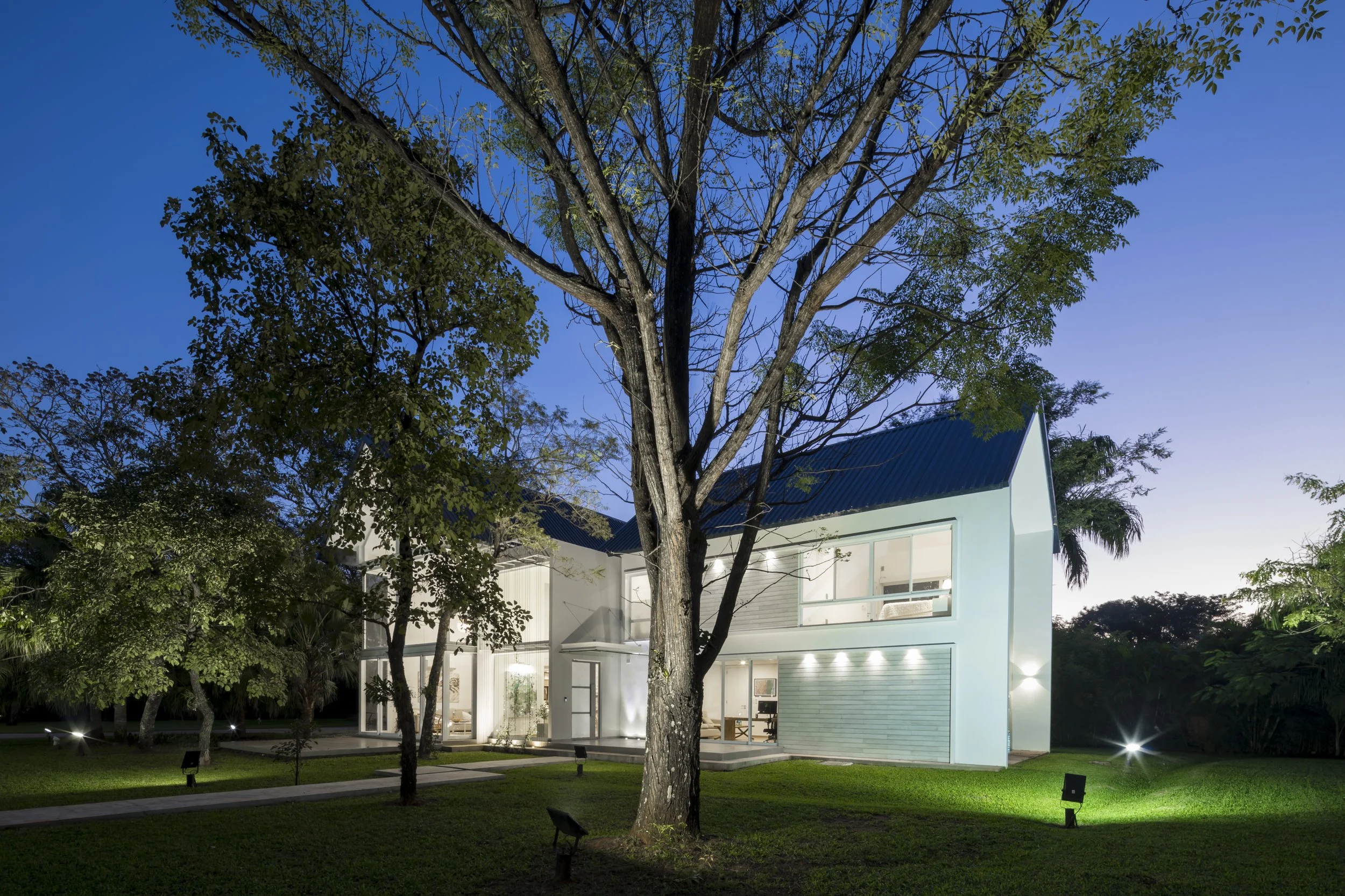 Casa moderna de dos pisos con paredes blancas y grande ventana, rodeada de árboles y césped, iluminada por luces exteriores en la noche.