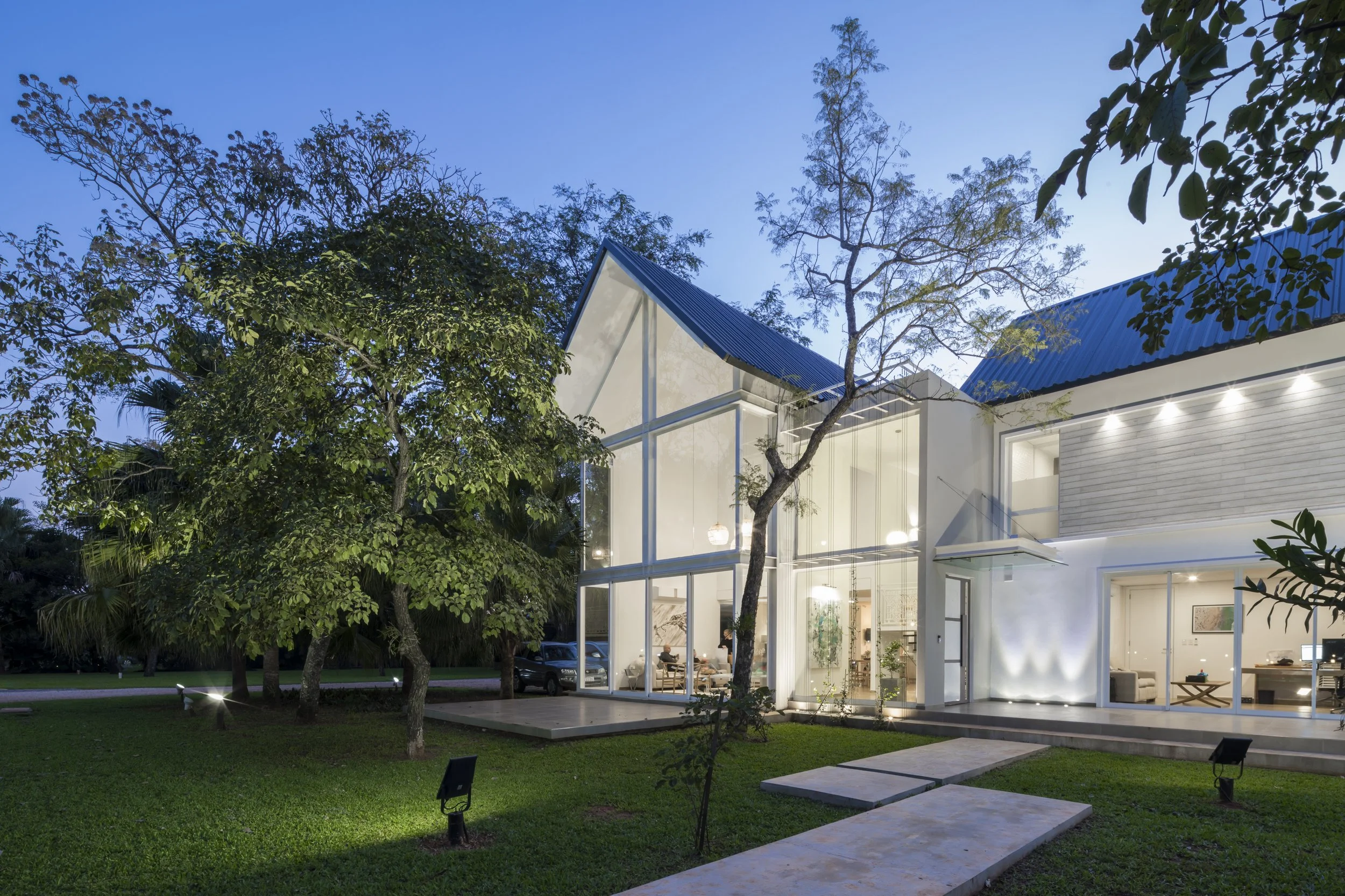 Casa moderna de dos pisos con paredes de vidrio y techo de lámina azul, rodeada de árboles y césped en la noche, iluminada por luces internas y externas.