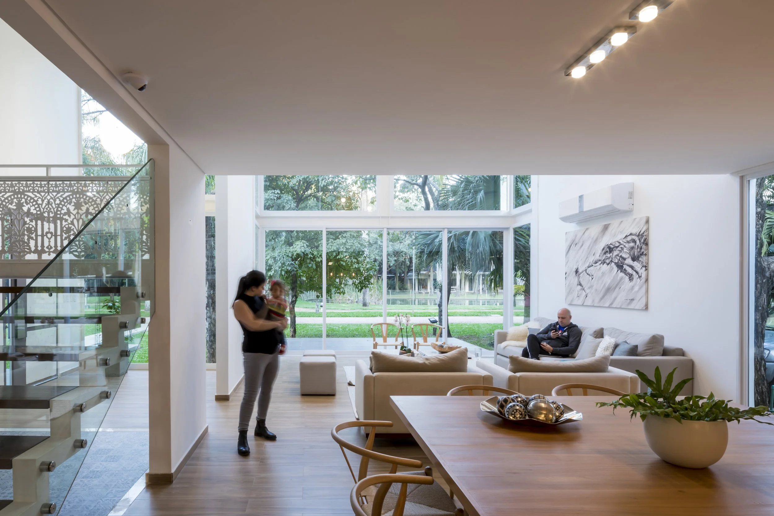 Sala moderna con ventanas grandes, mesa de madera con planta, sillones blancos, cuadro de pez en la pared, persona con perro en sillón, otra mujer con niño en área de sala, vista hacia un jardín exterior.