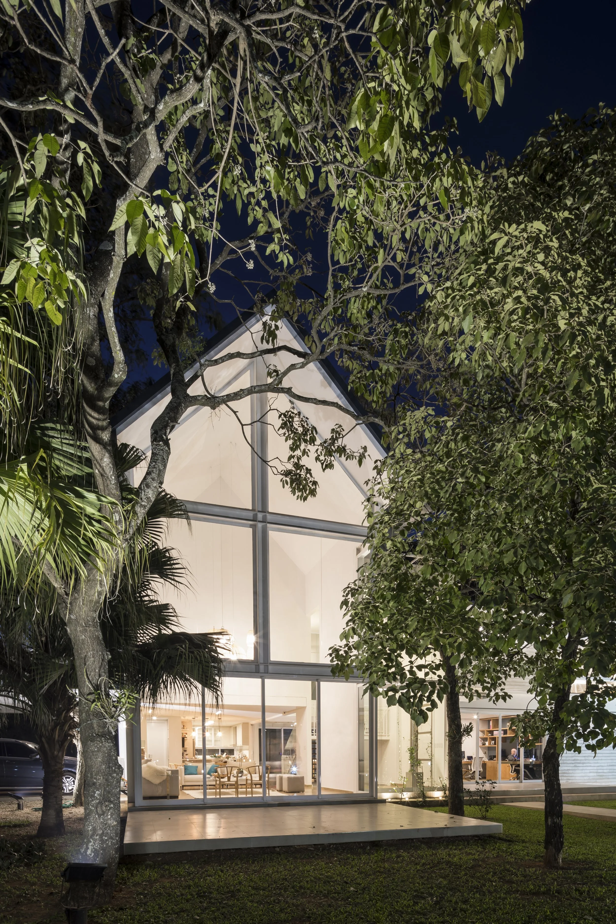 Casa moderna de dos pisos con grandes ventanales, iluminada en la noche, rodeada de árboles y césped.