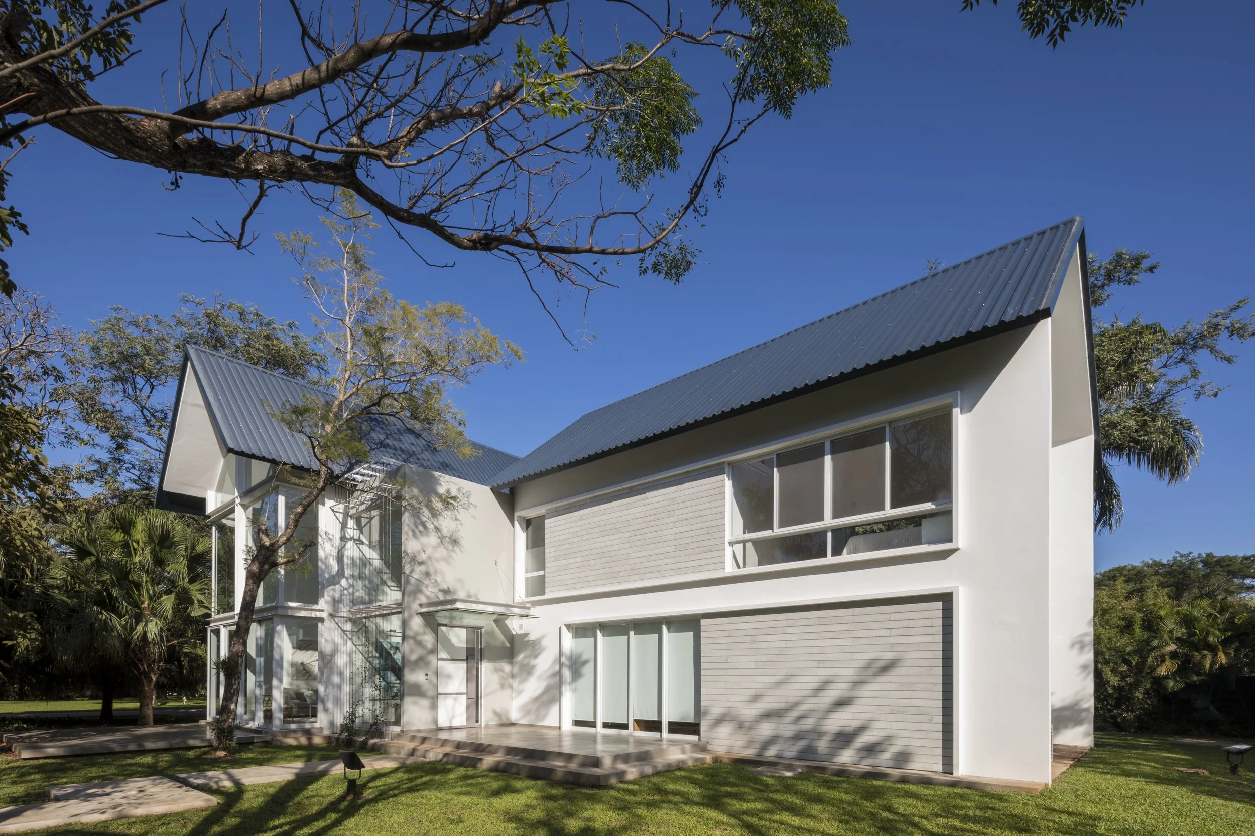 Casa moderna de dos pisos con paredes blancas, techos de metal y grandes ventanas, rodeada de árboles y césped bajo un cielo despejado.