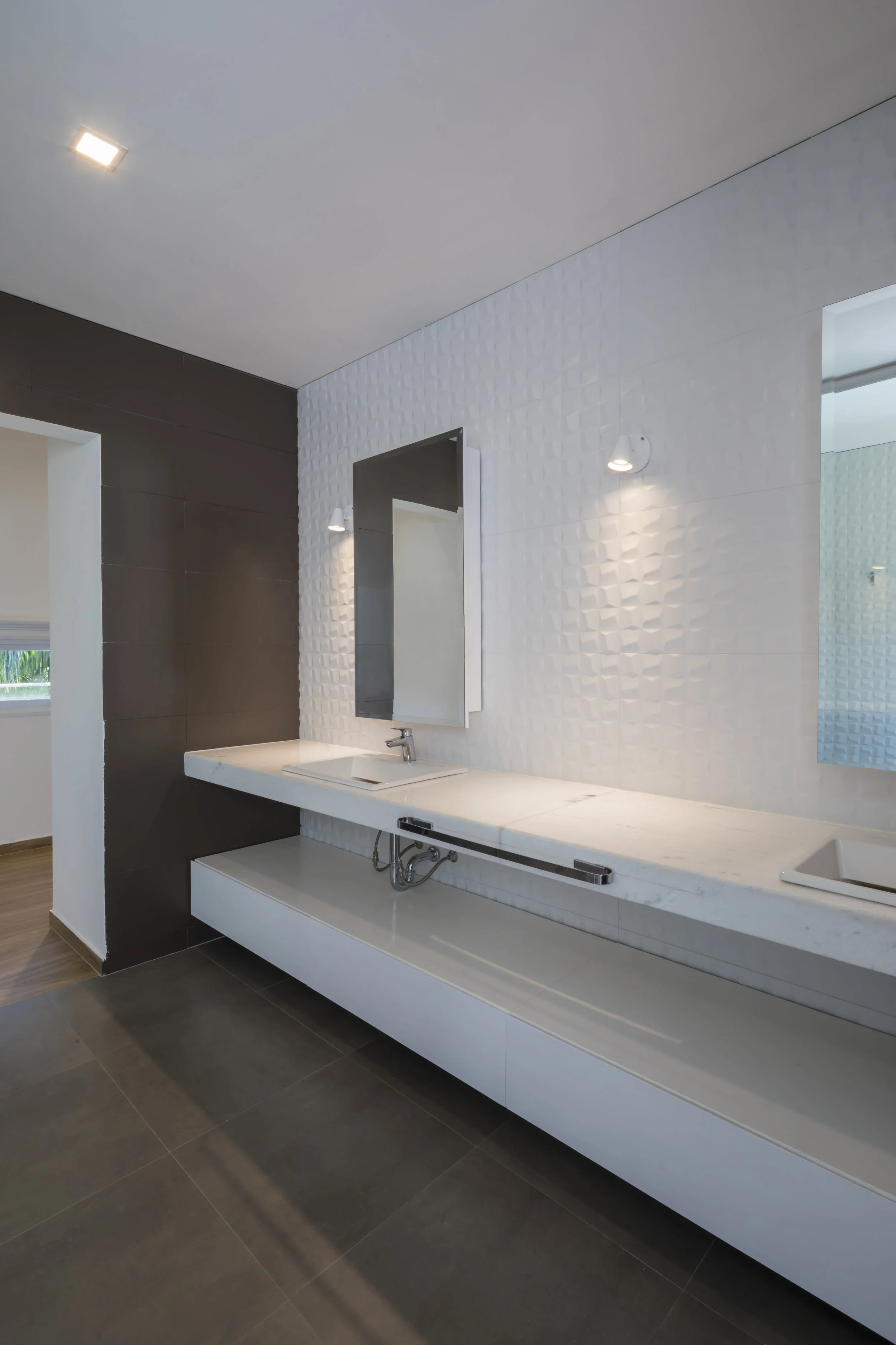 Baño moderno con pared blanca texturizada, espejo grande y lavabo de mármol con gabinetes blancos debajo.