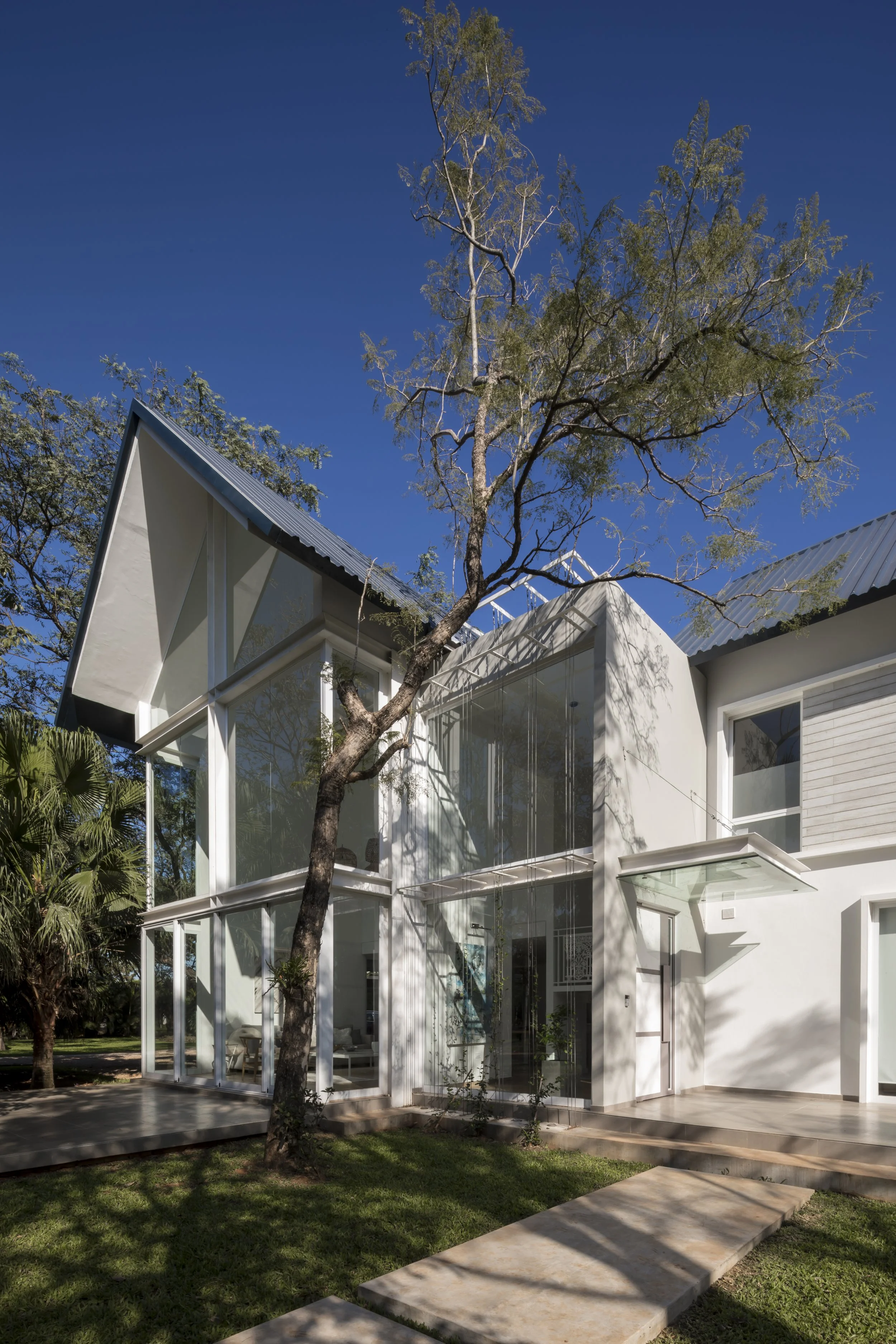 Casa moderna de dos pisos con paredes blancas, muchas ventanas de vidrio y techo de lámina metálica, rodeada de árboles y césped, en un día soleado con cielo azul.