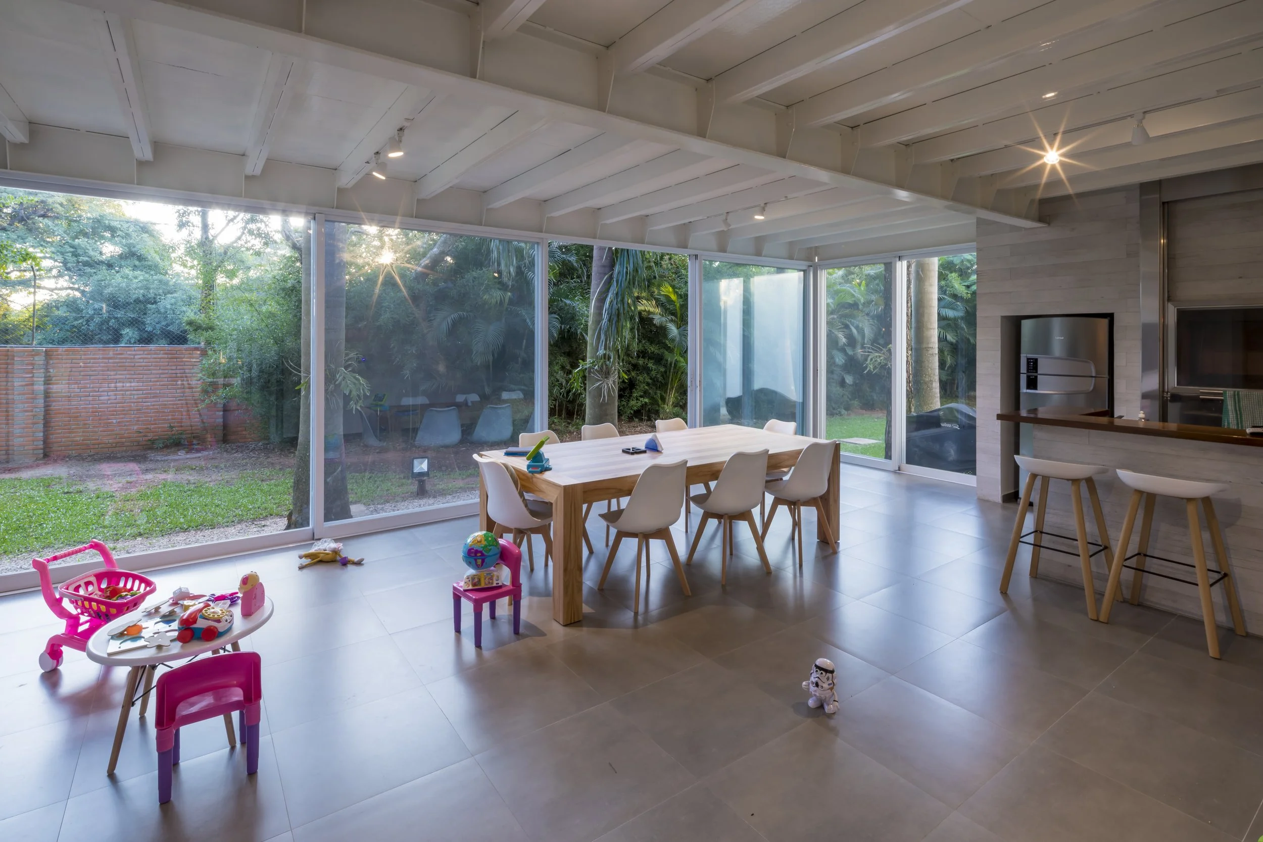 Comedor interior con mesa de madera y sillas blancas, juguetes de niños dispersos, ventanas grandes con vista a jardín con árboles y césped, iluminación moderna, espacio abierto y luminoso.