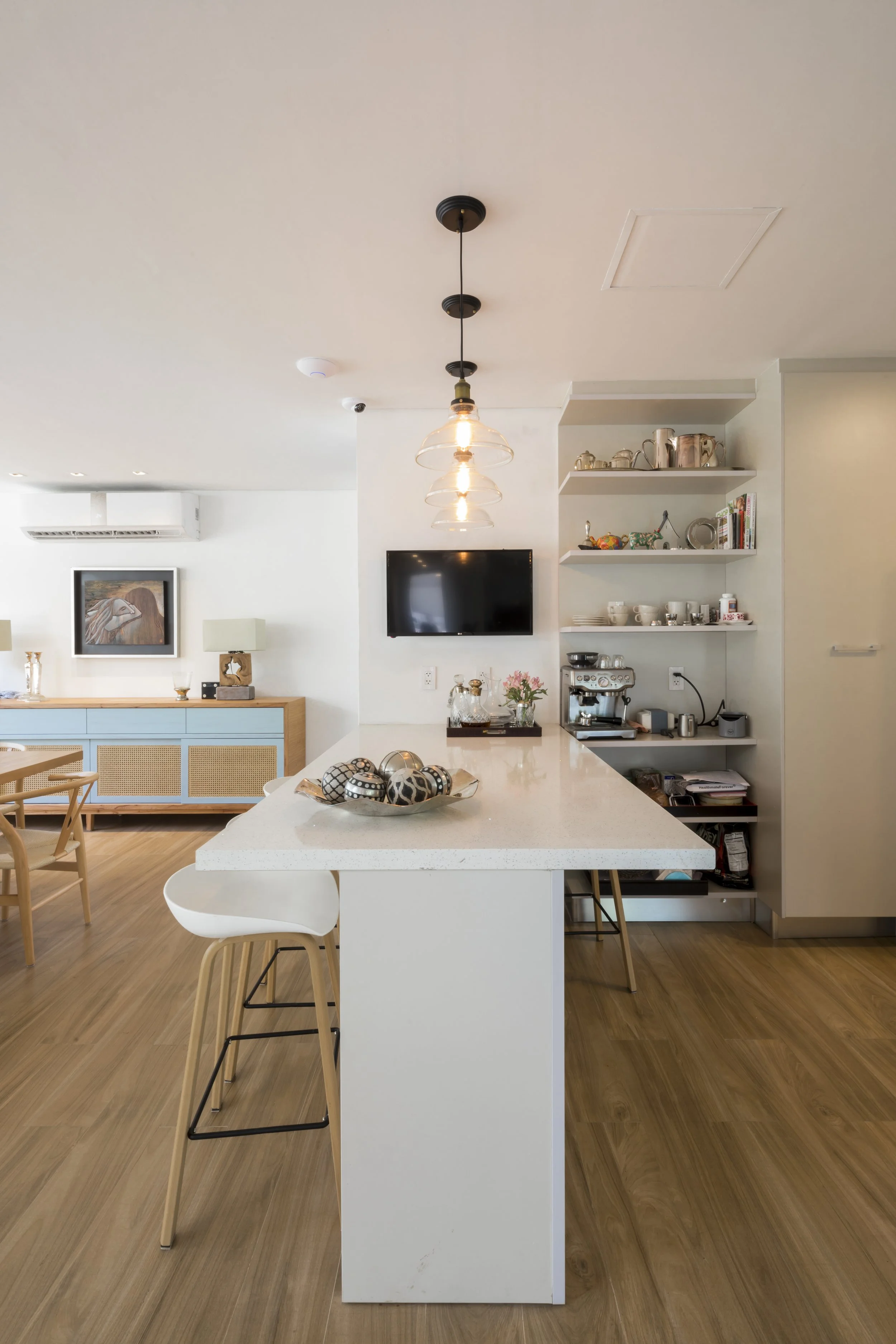Cocina moderna con isla blanca, barra con sillas blancas, estantes con vajilla y máquina de espresso, televisor en la pared, piso de madera.