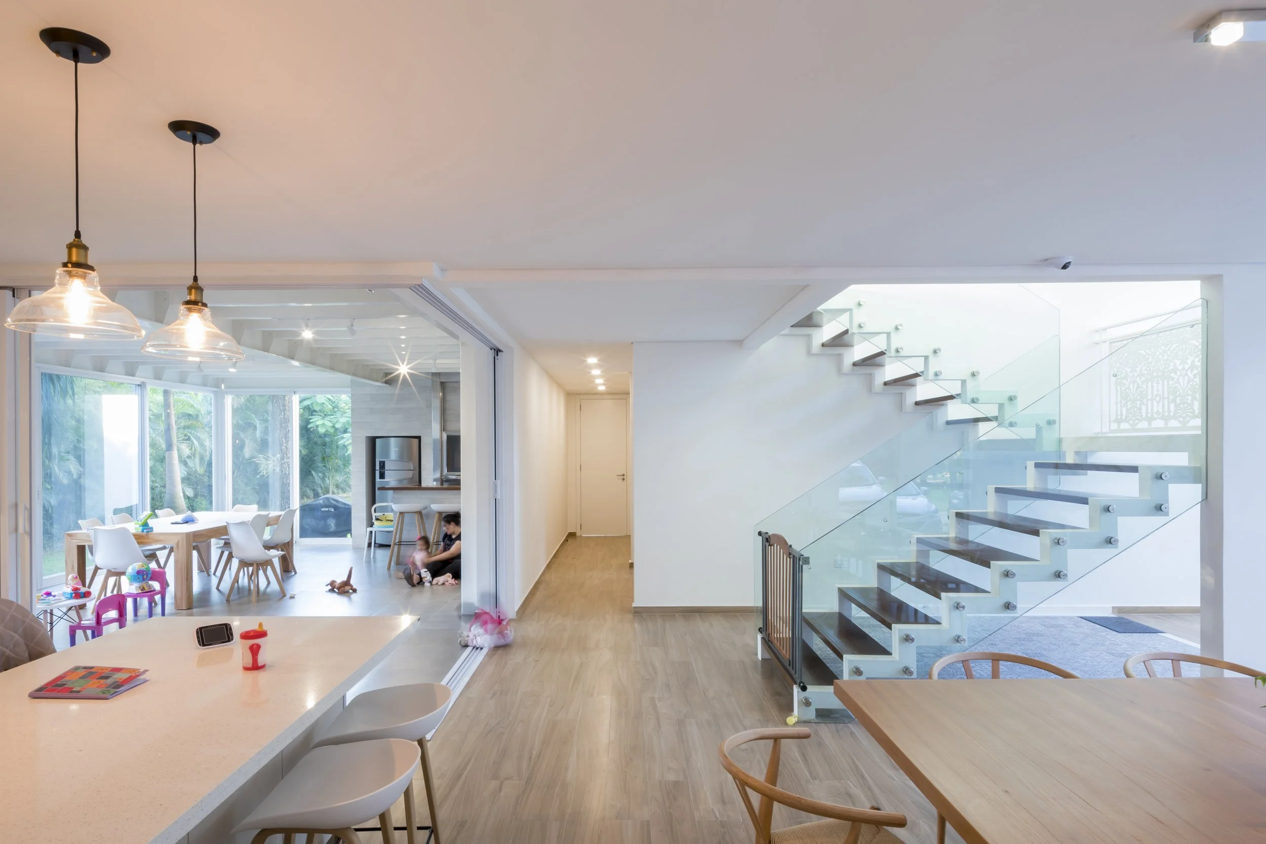 Interior de una casa moderna con cocina y sala, escaleras de vidrio, espacio bien iluminado y decorado con muebles de madera y sillas blancas.