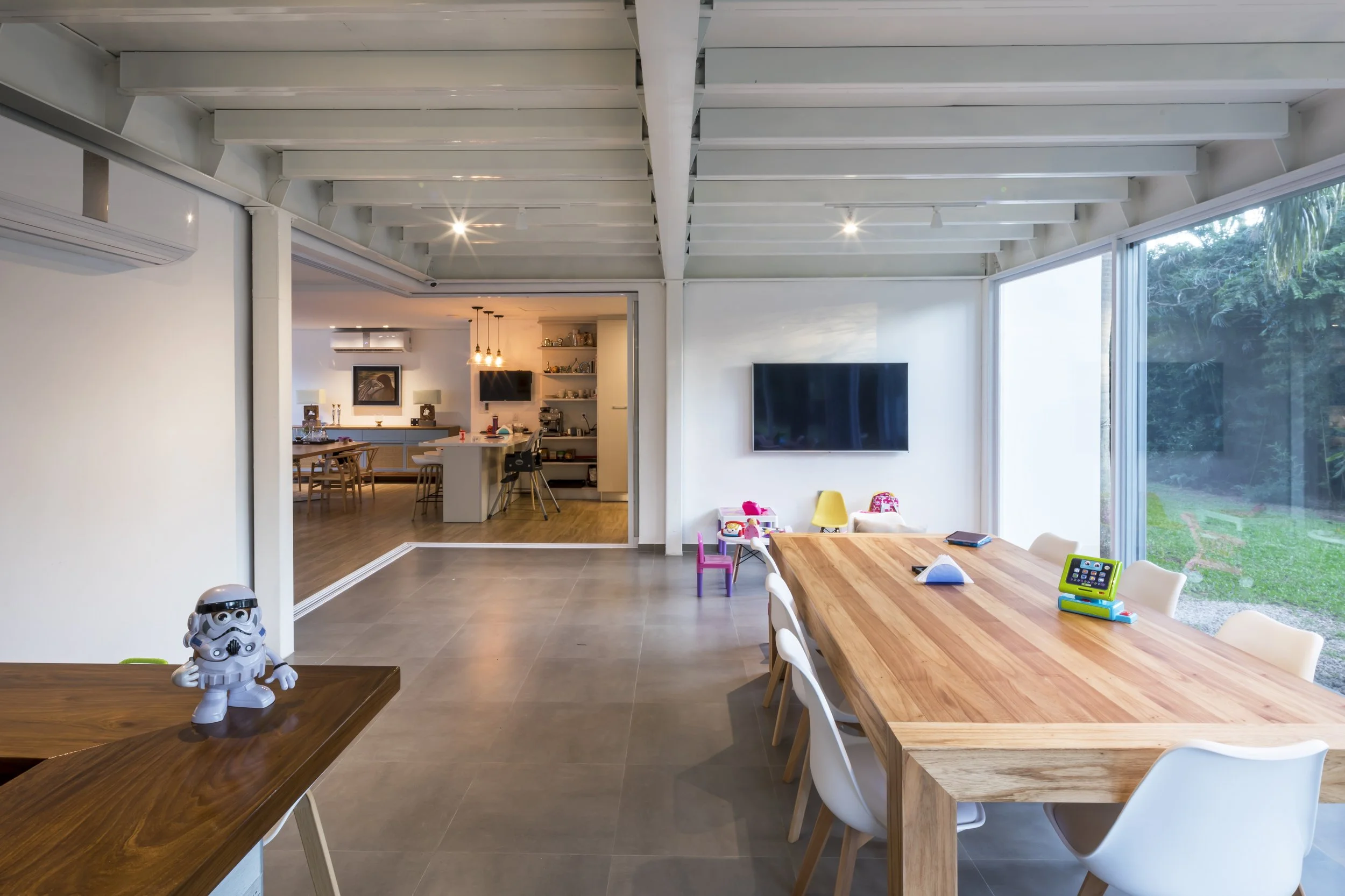 Sala de estar moderna con mesa grande de madera, sillas blancas, televisión en la pared, ventanas grandes con vista al jardín, juguetes para niños y una figura de un personaje de Star Wars en la mesa.
