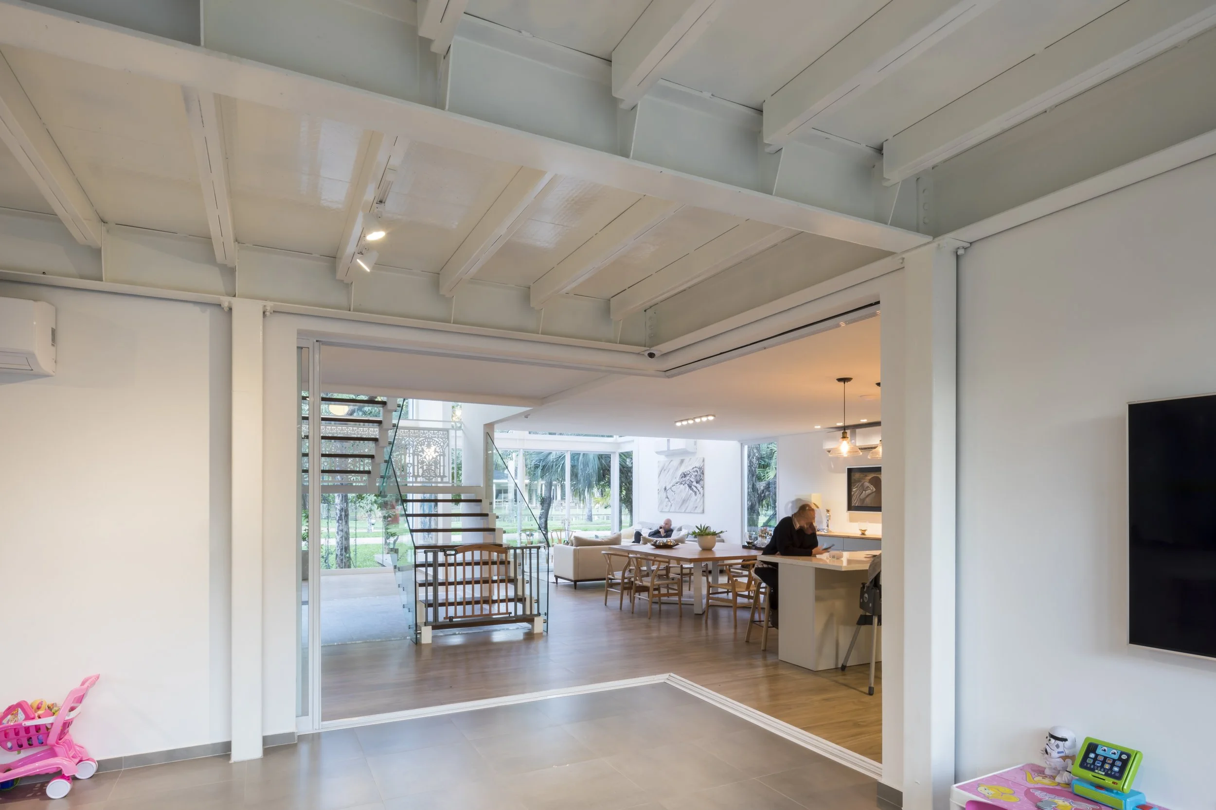 Vista interior de una sala moderna y luminosa con paredes blancas, pisos de madera, muchas ventanas grandes y un espacio abierto que conecta con un área de comedor y sala de estar.