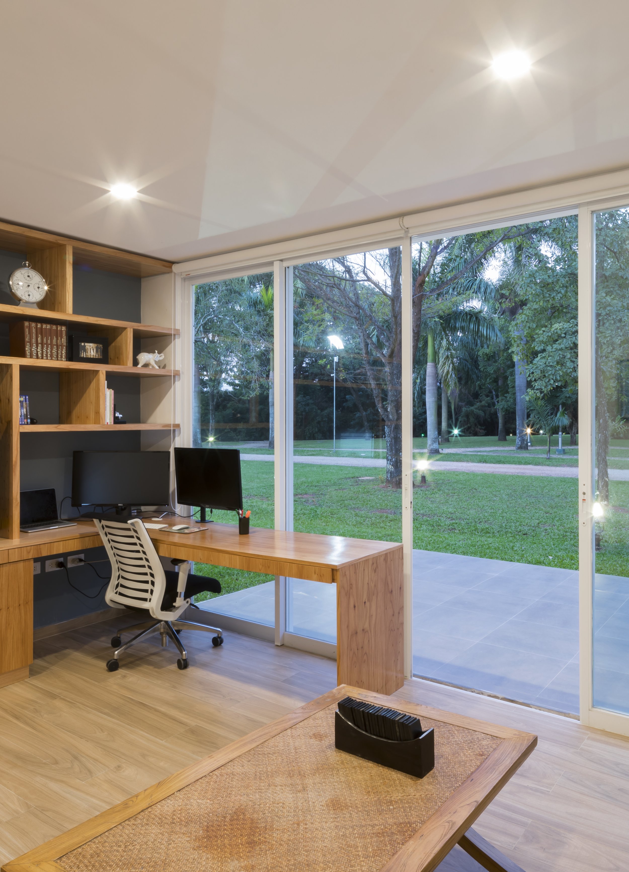 Oficina con vista a un jardín con árboles y césped, oficina con escritorio, computadora, estanterías y silla.