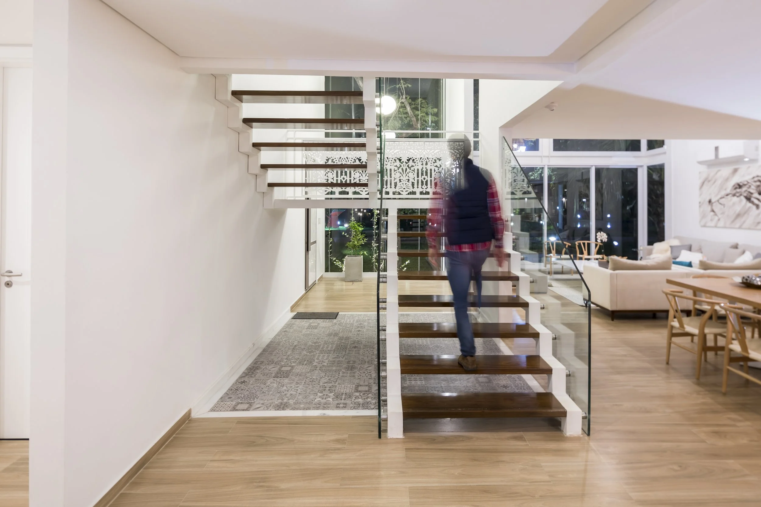 Interior moderno de casa, escalera de madera y vidrio, persona caminando, sala con sofá blanco y mesa de madera, grandes ventanas con vista al exterior, decoración minimalista