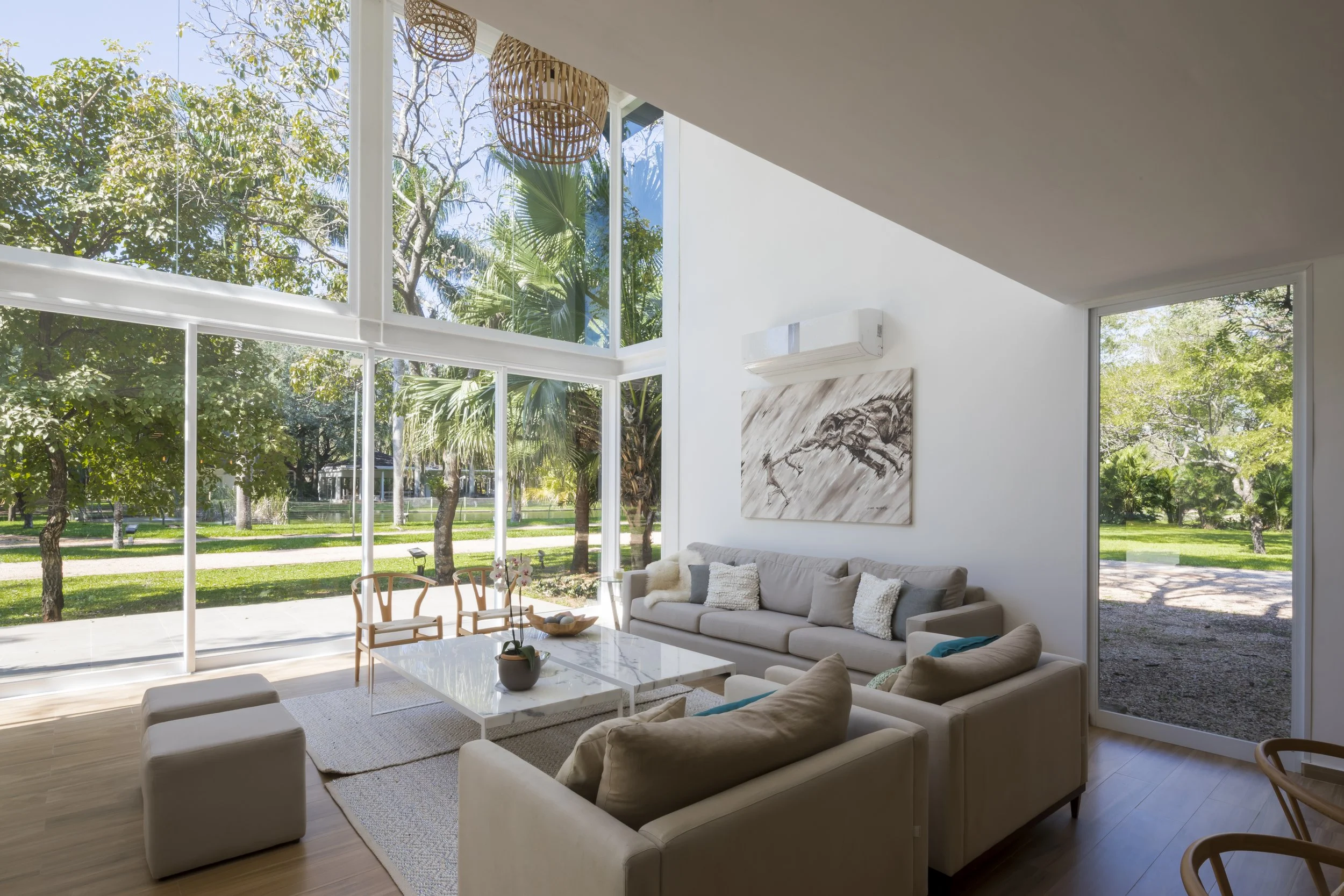 Interior de sala moderna con sillones beige, mesa de centro de mármol, pintura abstracta en la pared, gran ventanal con vista a jardín con árboles y césped, iluminación natural
