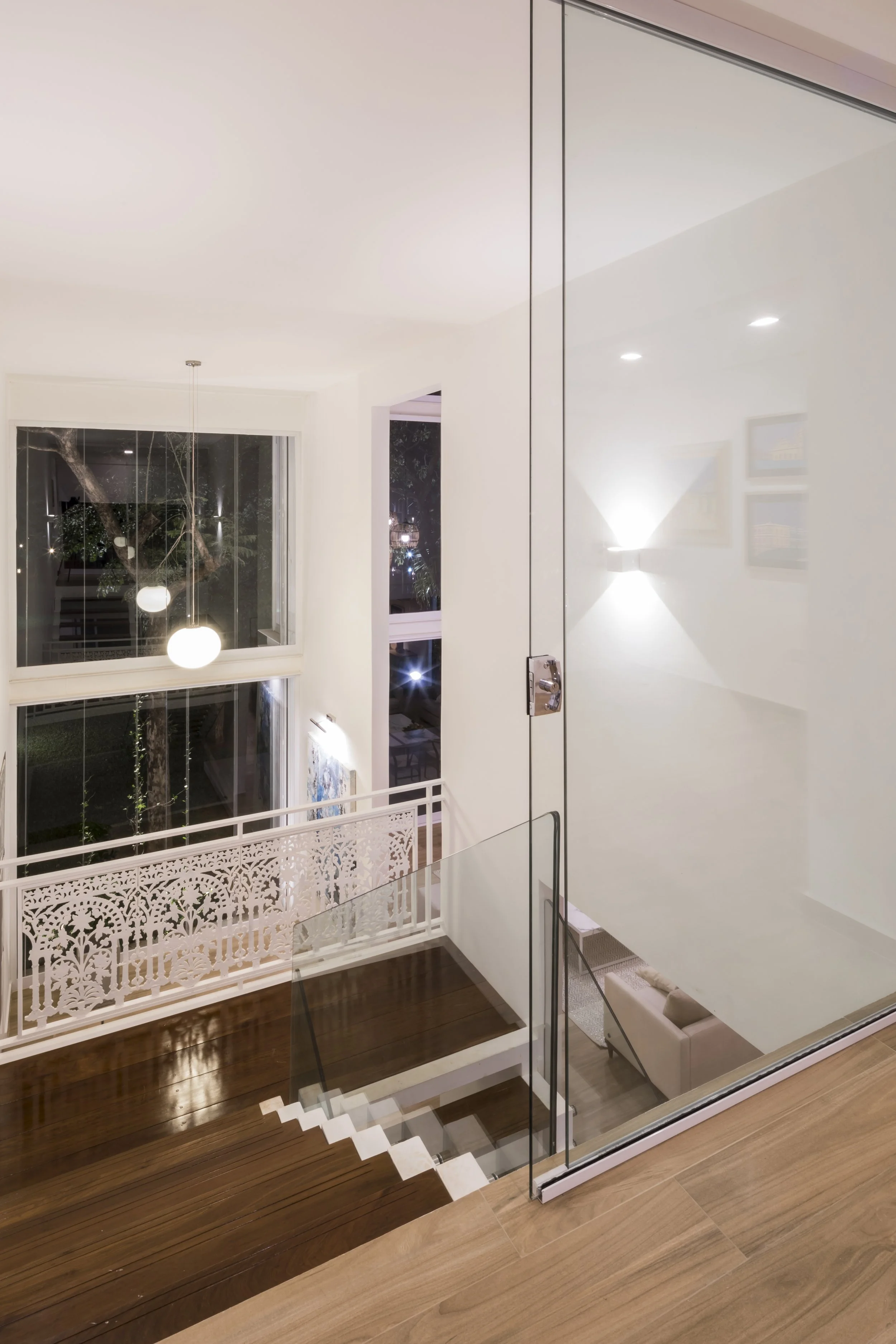 Vista interior de una casa moderna, con escalera de madera, barandales de vidrio y paredes blancas, en la noche con luz artificial encendida.