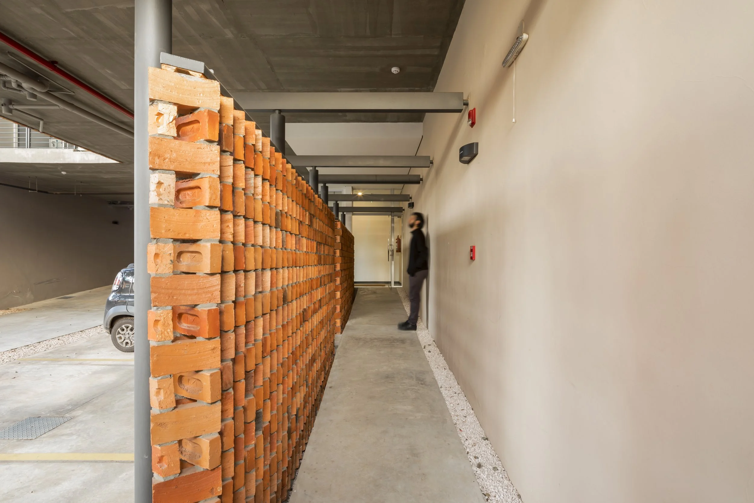 Pasillo en un edificio con pared de ladrillos a la izquierda y pared lisa a la derecha, con una persona de pie con máscara frente a la pared del lado derecho.