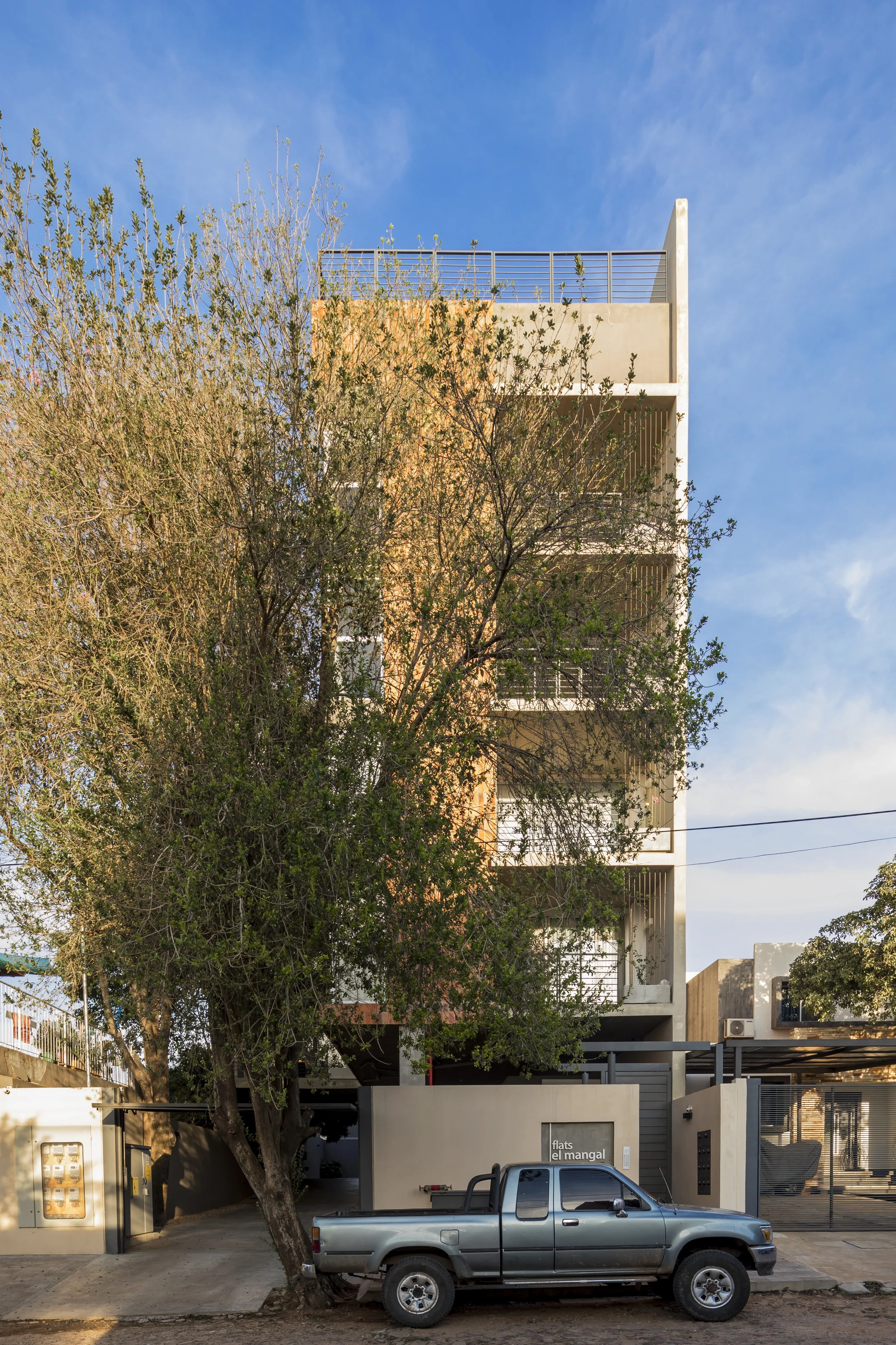Edificio residencial moderno de cuatro pisos con balcones, un árbol verde en primer plano y un camión gris estacionado en la calle. El cielo es despejado y azul.