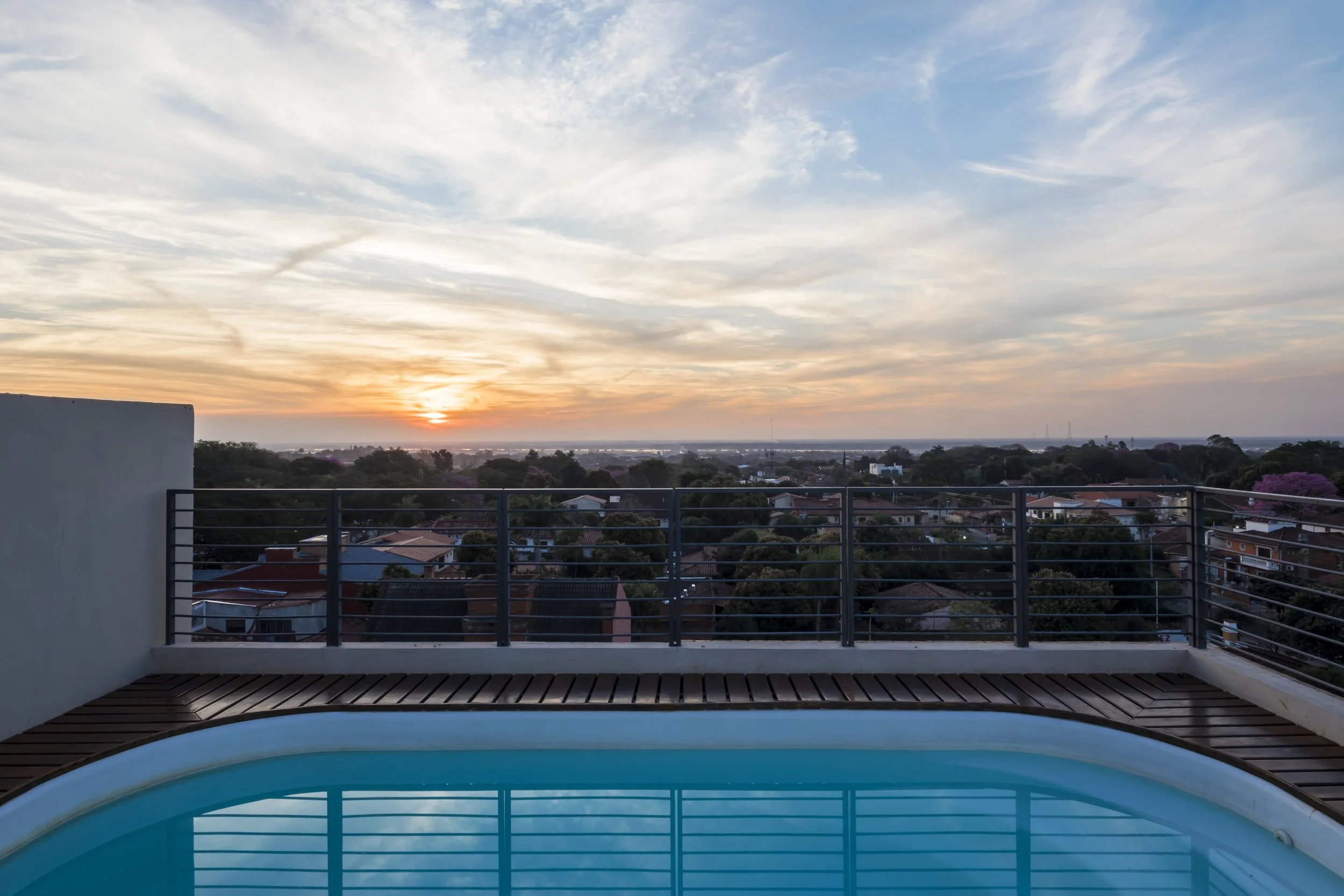 Vista del atardecer desde una azotea con una piscina en primer plano y un cielo con nubes y el sol bajando en el horizonte.