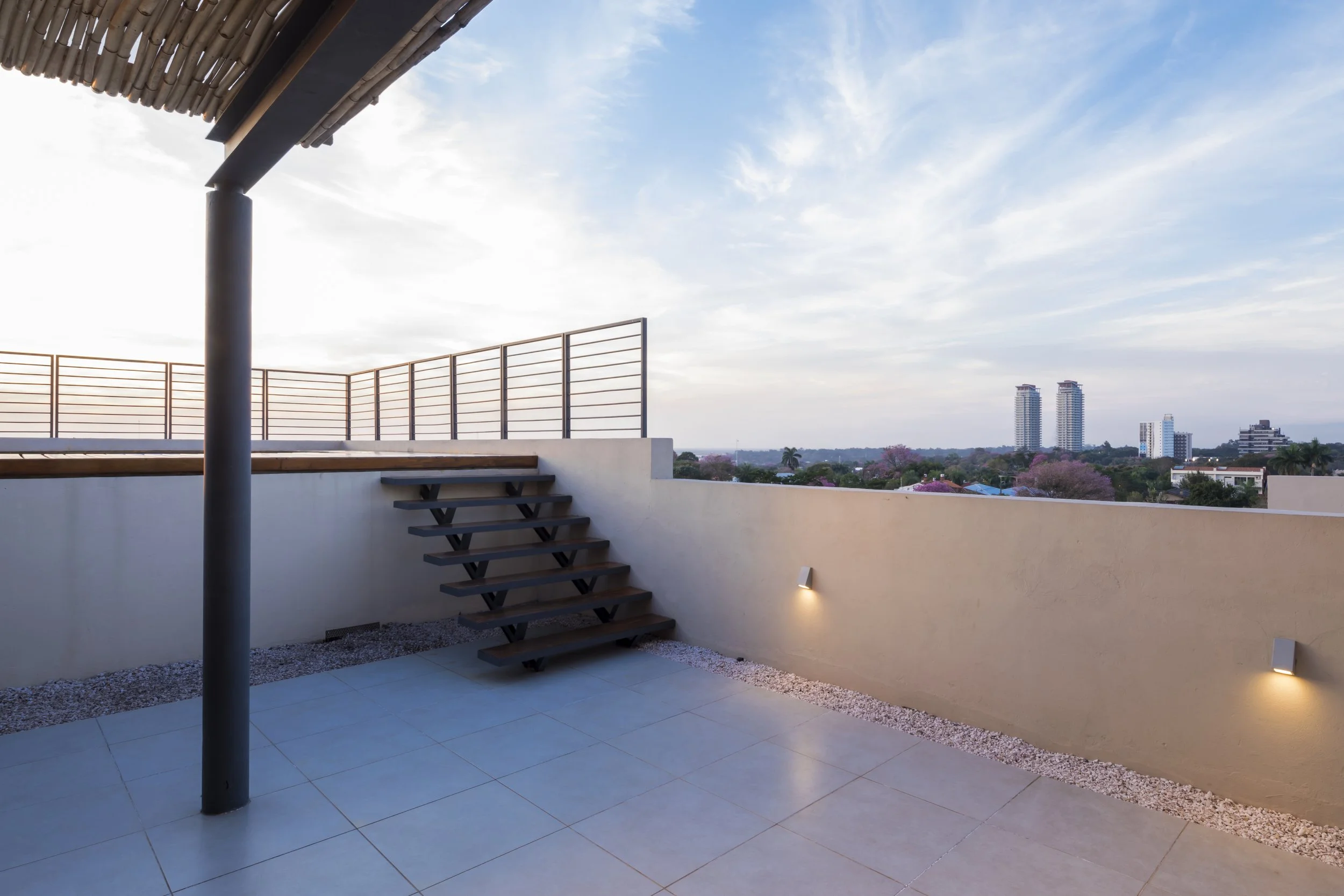Terraza con vista a la ciudad, suelo de baldosas, pared beige con luces empotradas, escaleras de madera y estructura de techumbre de madera.