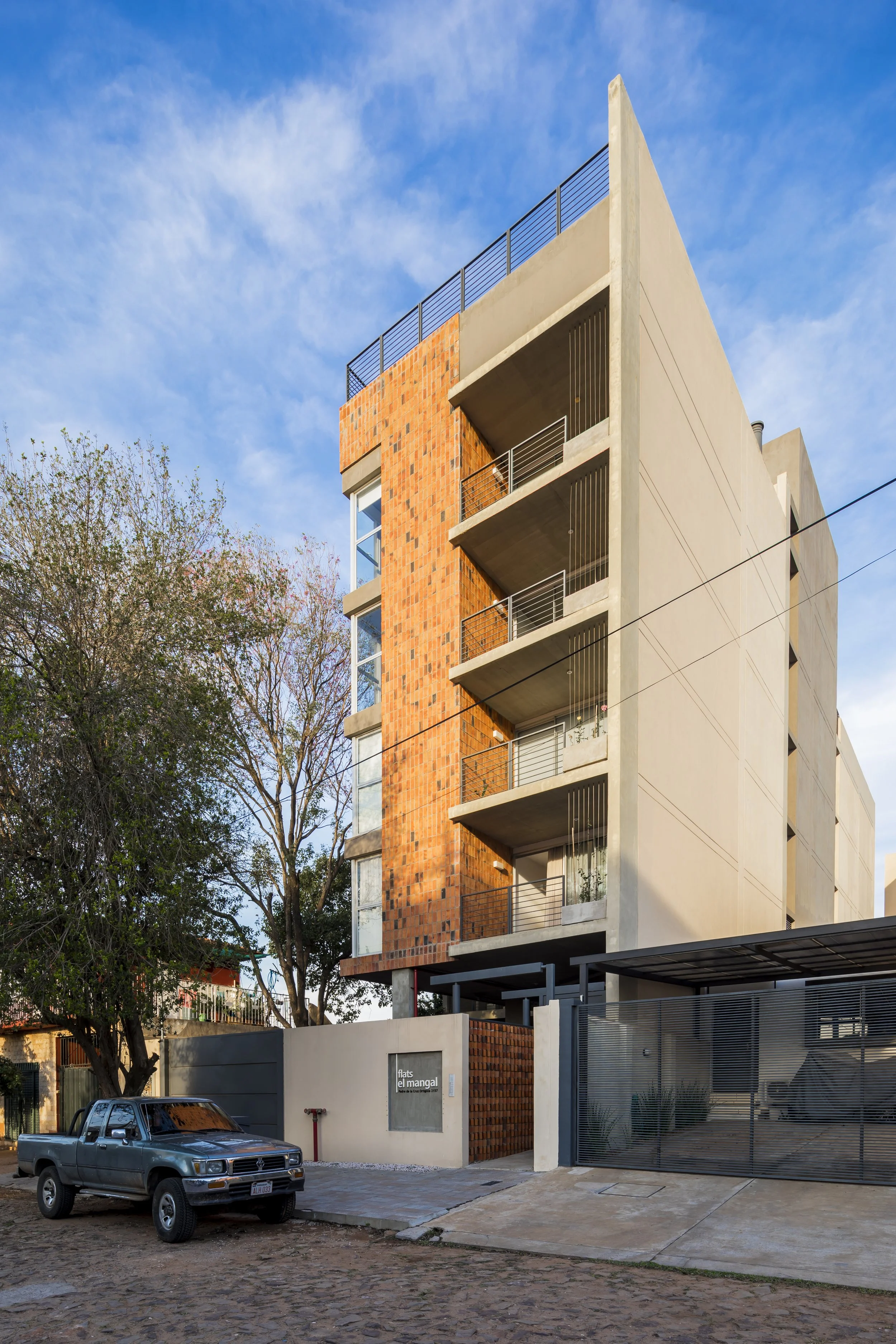 Edificio moderno de varios pisos con balcones, fachada de ladrillos y concreto en una calle con árbol y carro frente.
