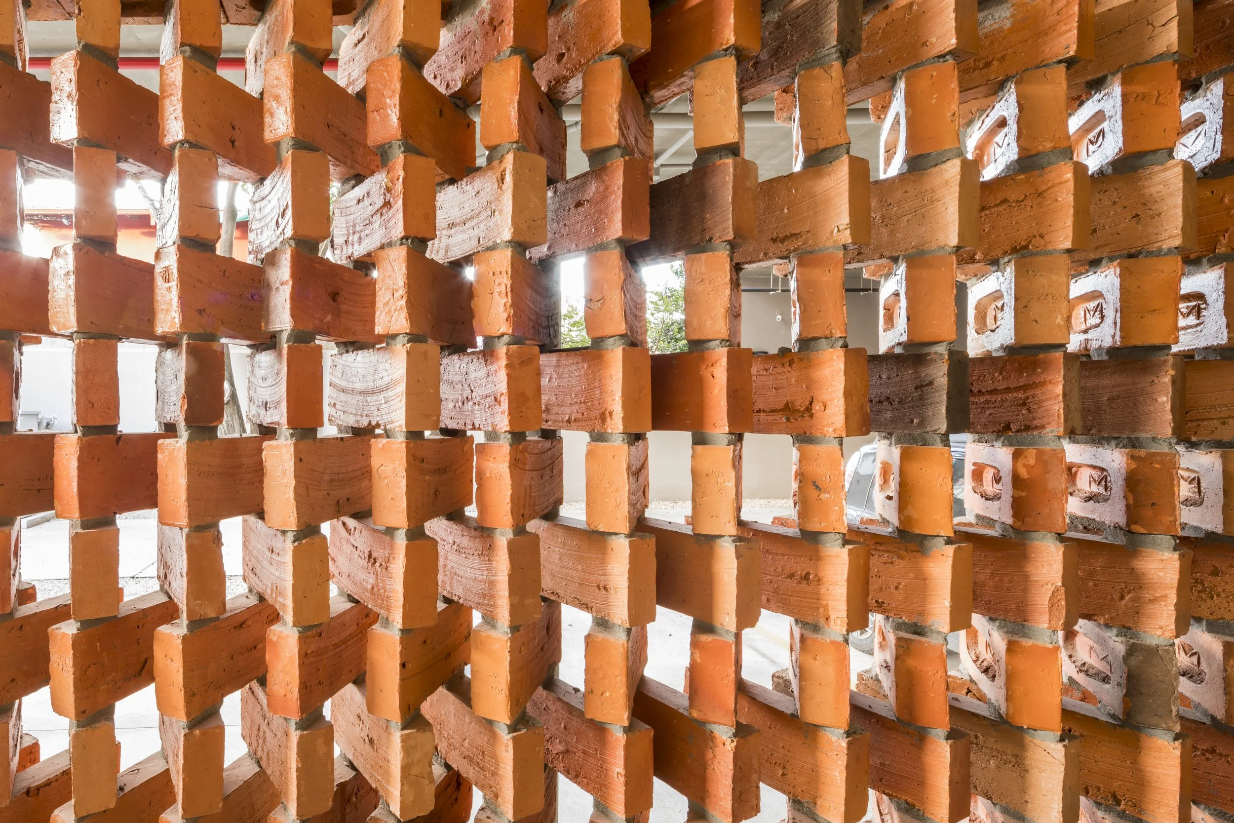 PRO ARQUITECTURA PARAGUAY architecture design PROSA
Muro de ladrillos de color rojo con espacios vacíos en el centro, vista desde el interior hacia afuera, con vehículos y árboles visibles al fondo.