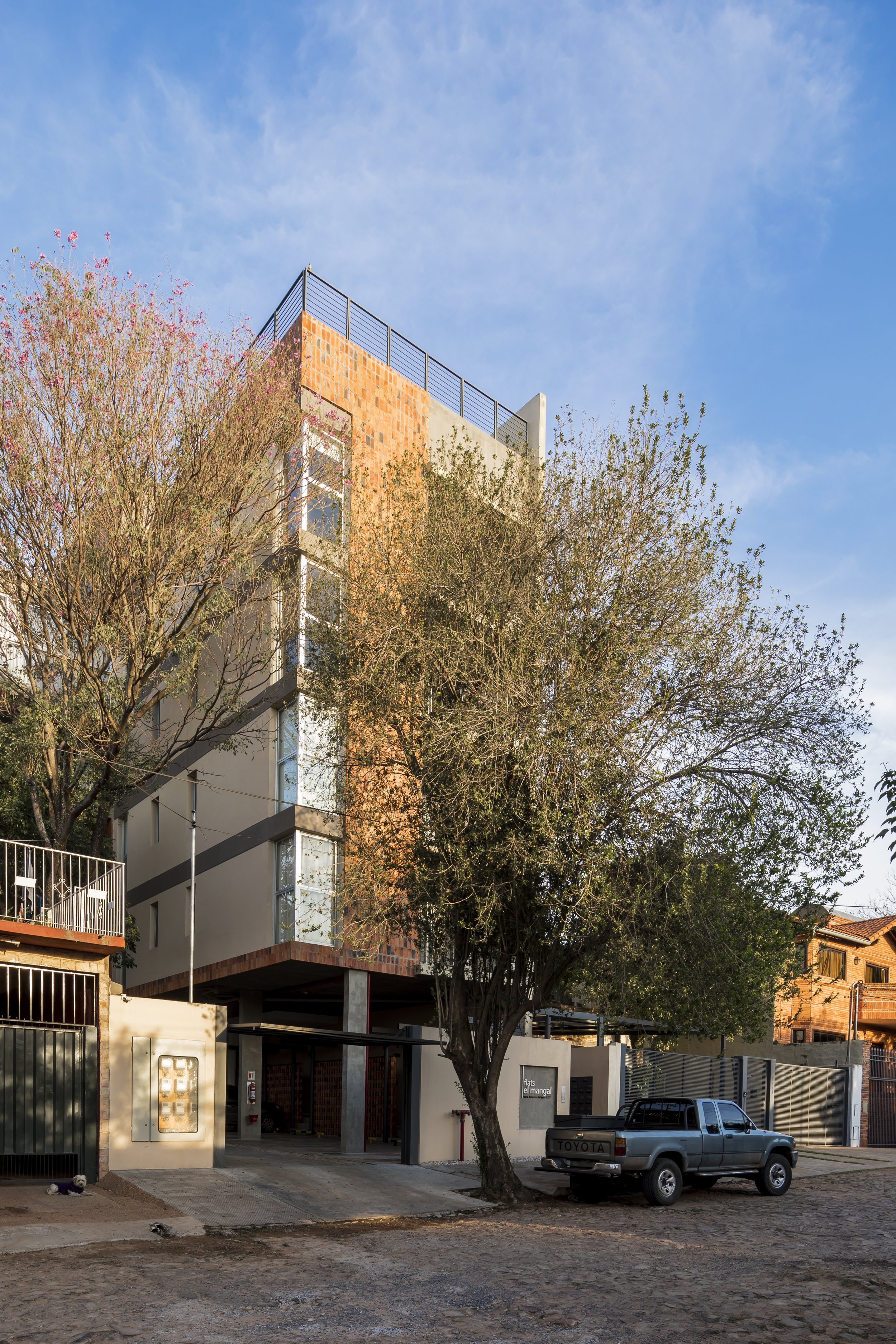 Edificio moderno de varios pisos con árboles y un coche Toyota estacionado en la calle, bajo un cielo despejado.