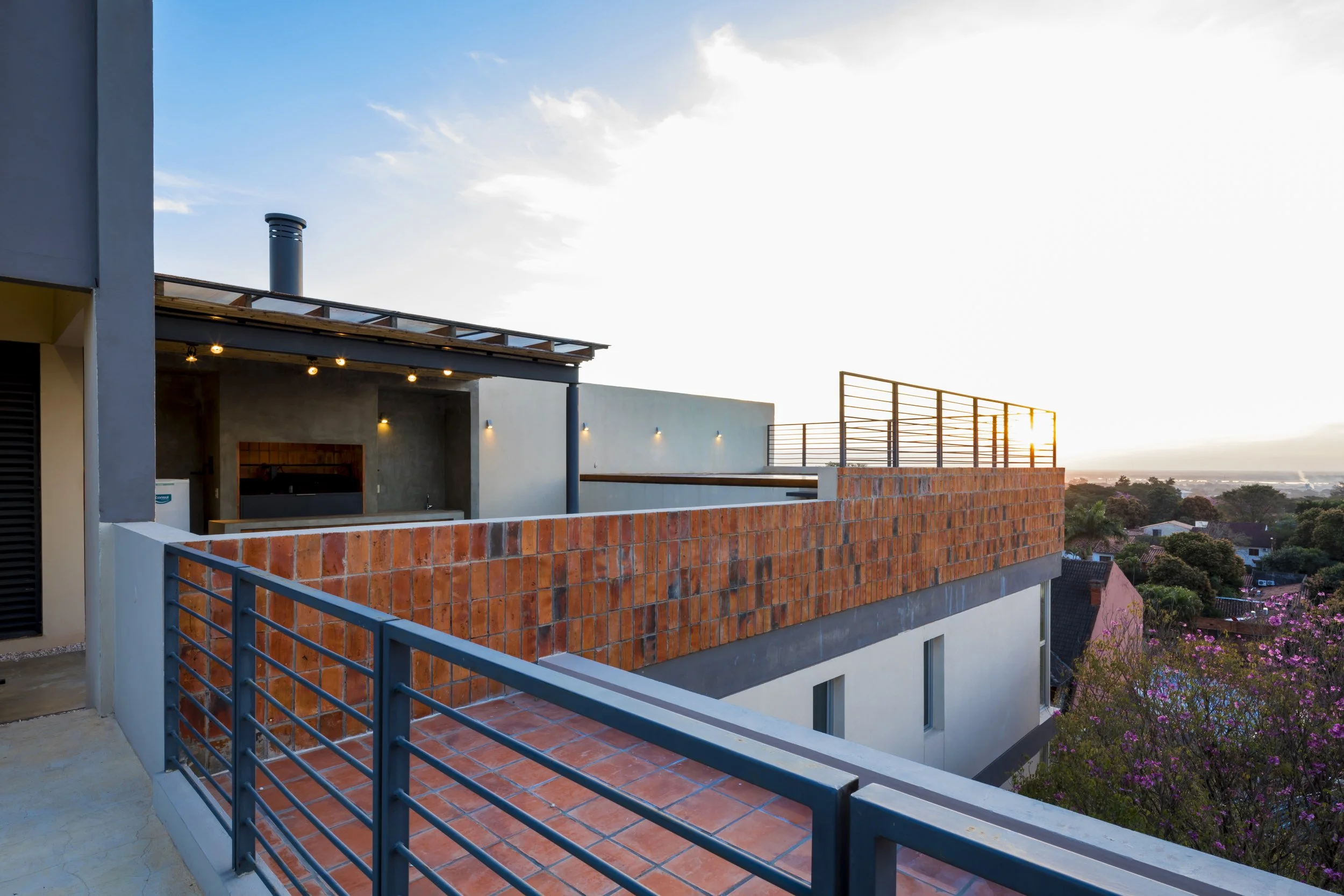 Vista de una terraza en un edificio moderno al atardecer, con barandillas de metal, paredes de ladrillo y una vista panorámica de casas y árboles en el fondo.