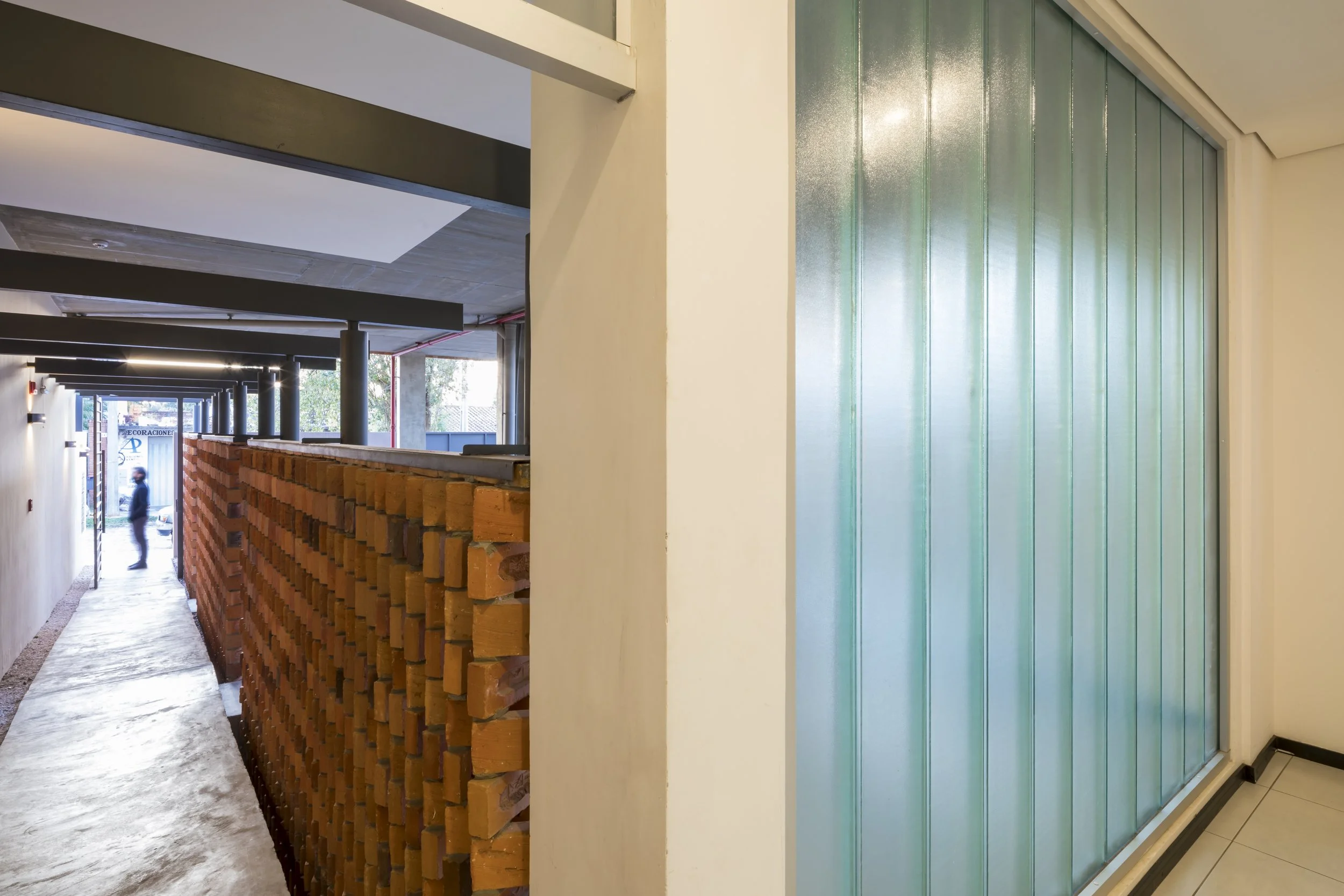 Pasillo en construcción con pared de ladrillos y estructura de techo, vista desde el interior de un edificio, con una puerta de vidrio opaca a la derecha.
