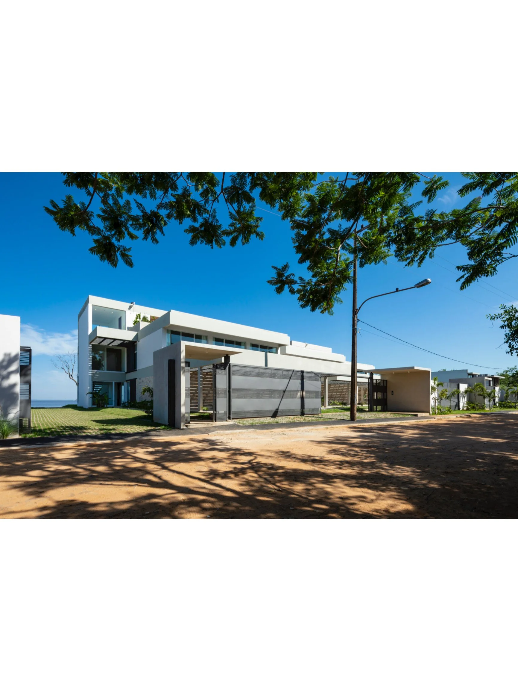 PRO ARQUITECTURA PARAGUAY architecture design PROSA
Casa moderna blanca con jardín y sombra de árboles, en una calle con poste de luz y cielo despejado.