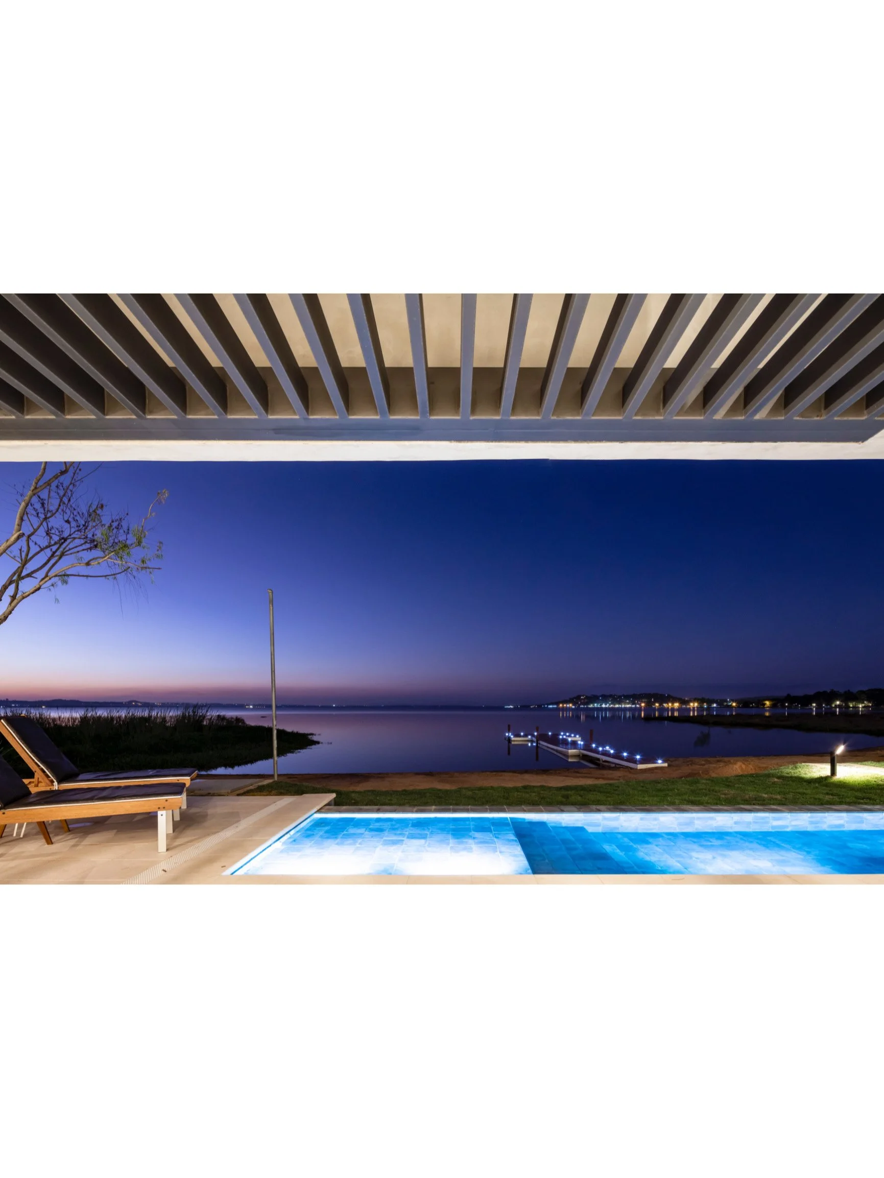 PRO ARQUITECTURA PARAGUAY architecture design PROSA
Vista nocturna de una piscina iluminada junto a un lago o río, con un muelle y luces en la orilla, bajo un cielo oscuro con un toque de luz en el horizonte.