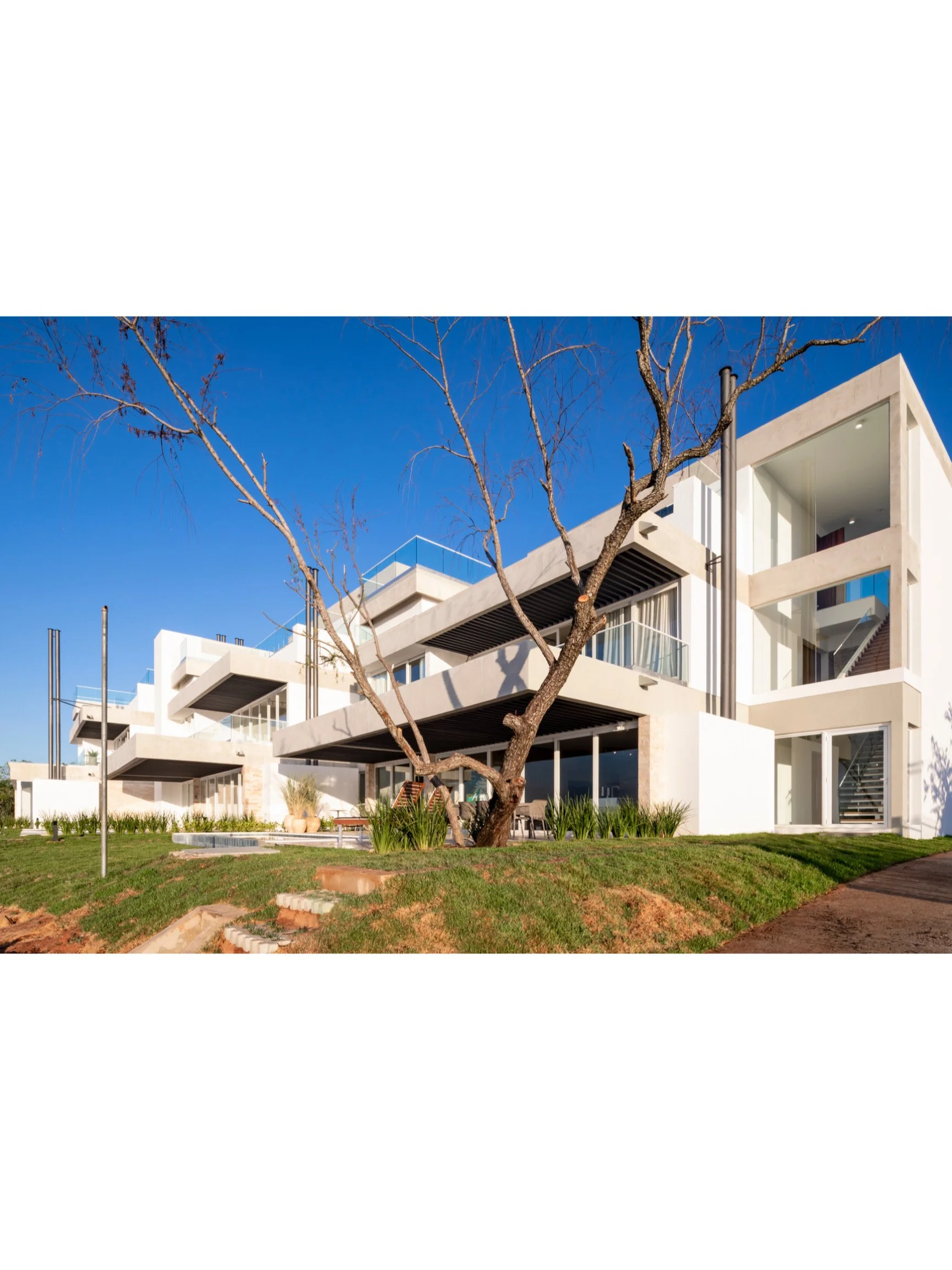 PRO ARQUITECTURA PARAGUAY architecture design PROSA
Edificio moderno de varios pisos con arquitectura blanca, grandes ventanas, balcón y escaleras en el exterior, con árbol sin hojas en primer plano y césped en la base, bajo cielo azul claro.