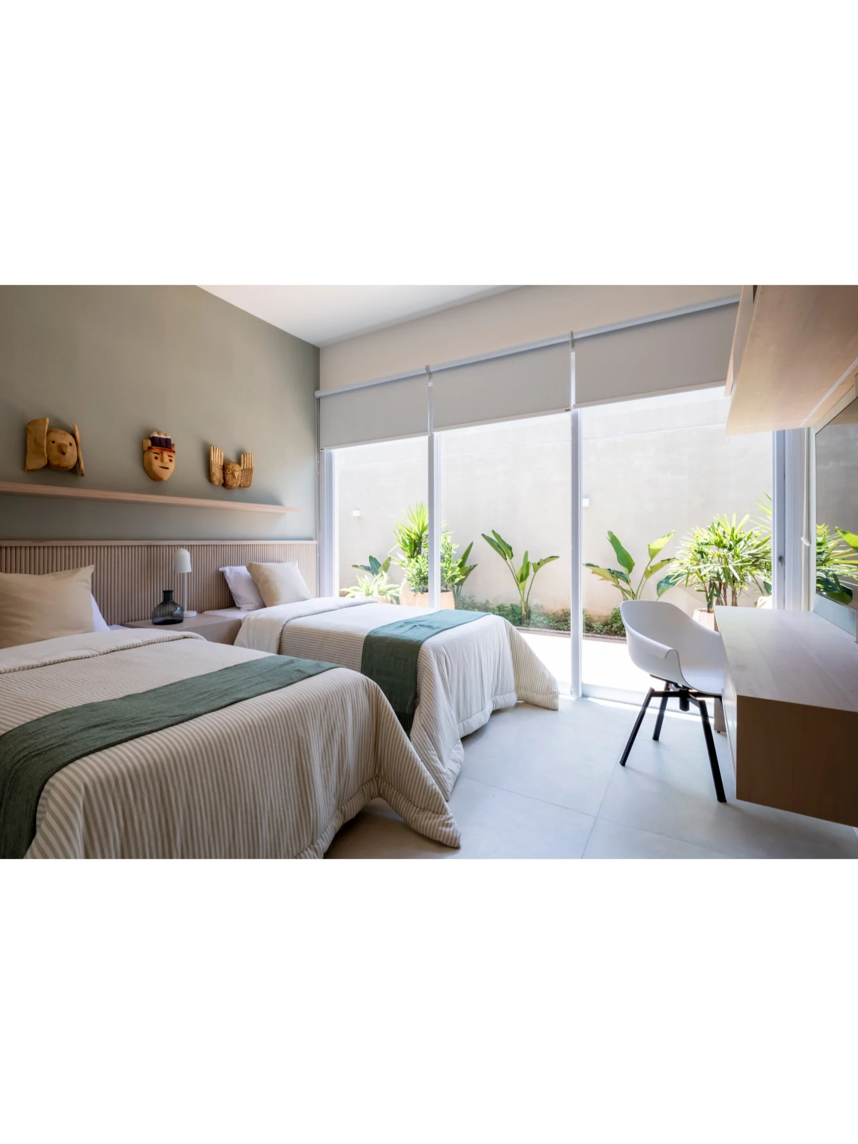 PRO ARQUITECTURA PARAGUAY architecture design PROSA
Habitación con dos camas, vista a un espacio exterior con plantas verdes, ventana grande con cortinas blancas, escritorio con silla, y decoraciones de máscaras en la pared.