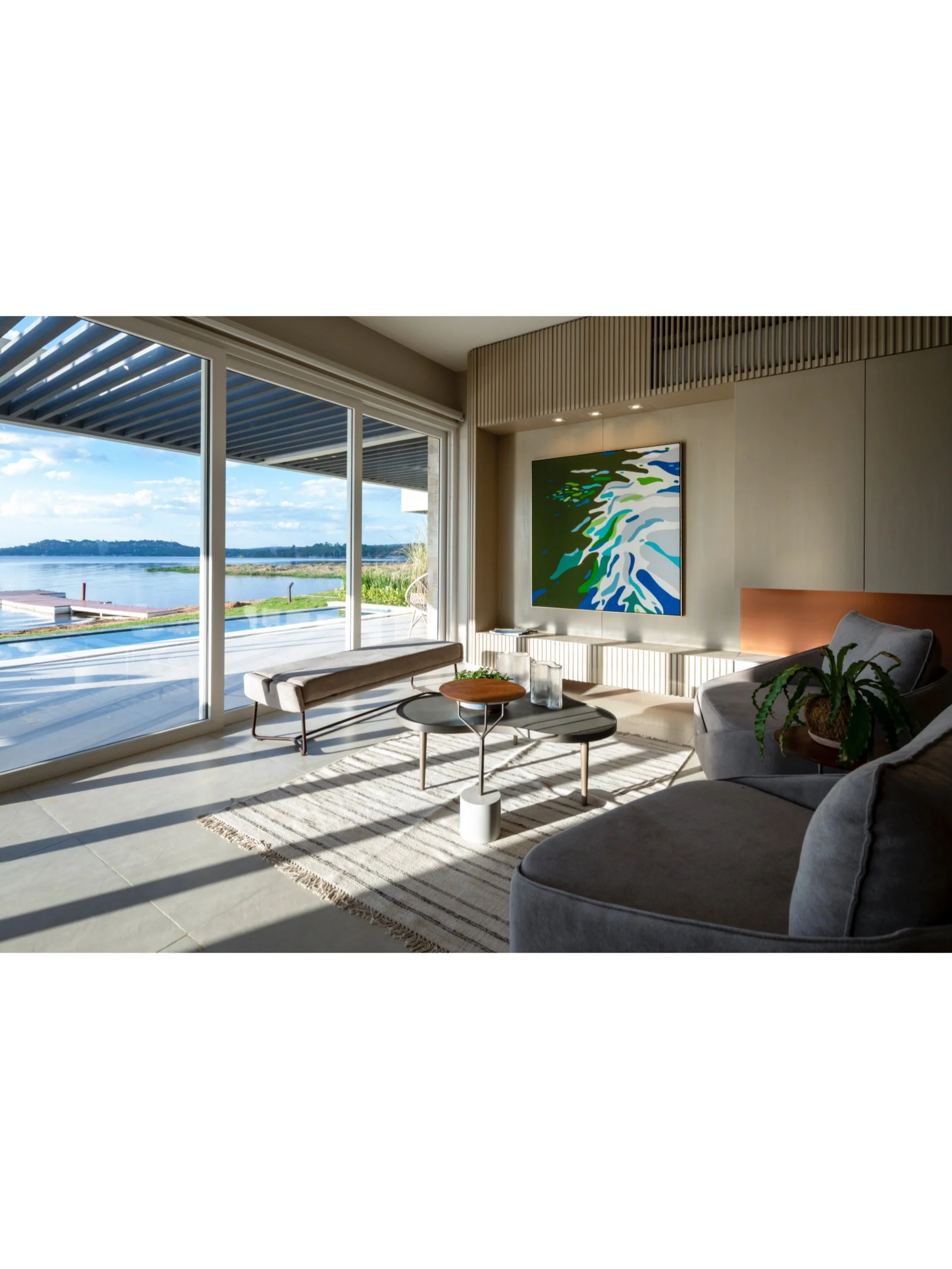 PRO ARQUITECTURA PARAGUAY architecture design PROSA
Sala de estar con vista al río, ventanas grandes, sillón gris, mesa de centro, obra de arte abstracta en la pared, plantas y decoración moderna.