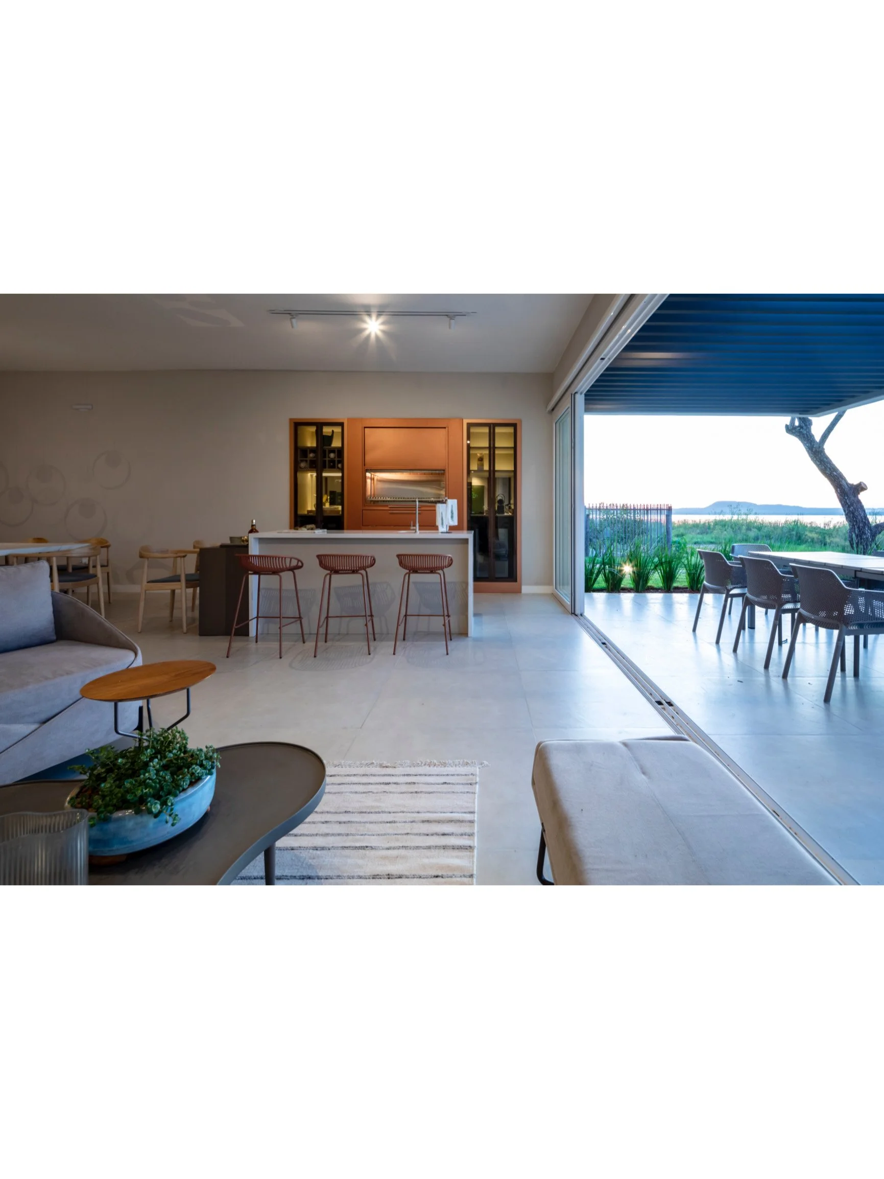 PRO ARQUITECTURA PARAGUAY architecture design PROSA
Interior de una sala de estar moderna con muebles de color neutro, una cocina con barra y sillas, y una terraza exterior con mesas y sillas, junto a un paisaje natural con árboles y agua.