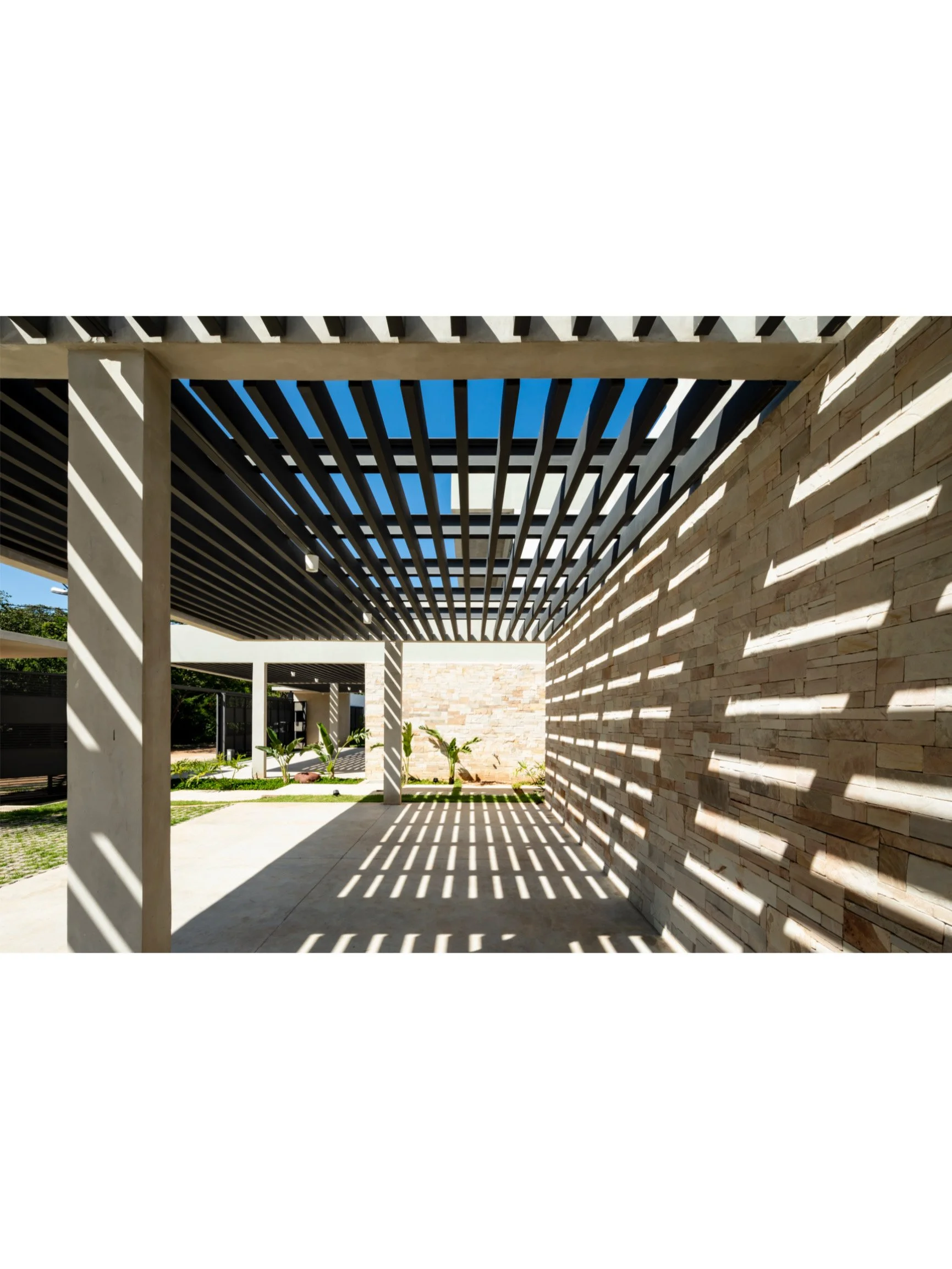 PRO ARQUITECTURA PARAGUAY architecture design PROSA
Perspectiva de un espacio exterior con estructura de vigas en el techo que proyecta sombras en la pared de bloques de piedra, en un día soleado.