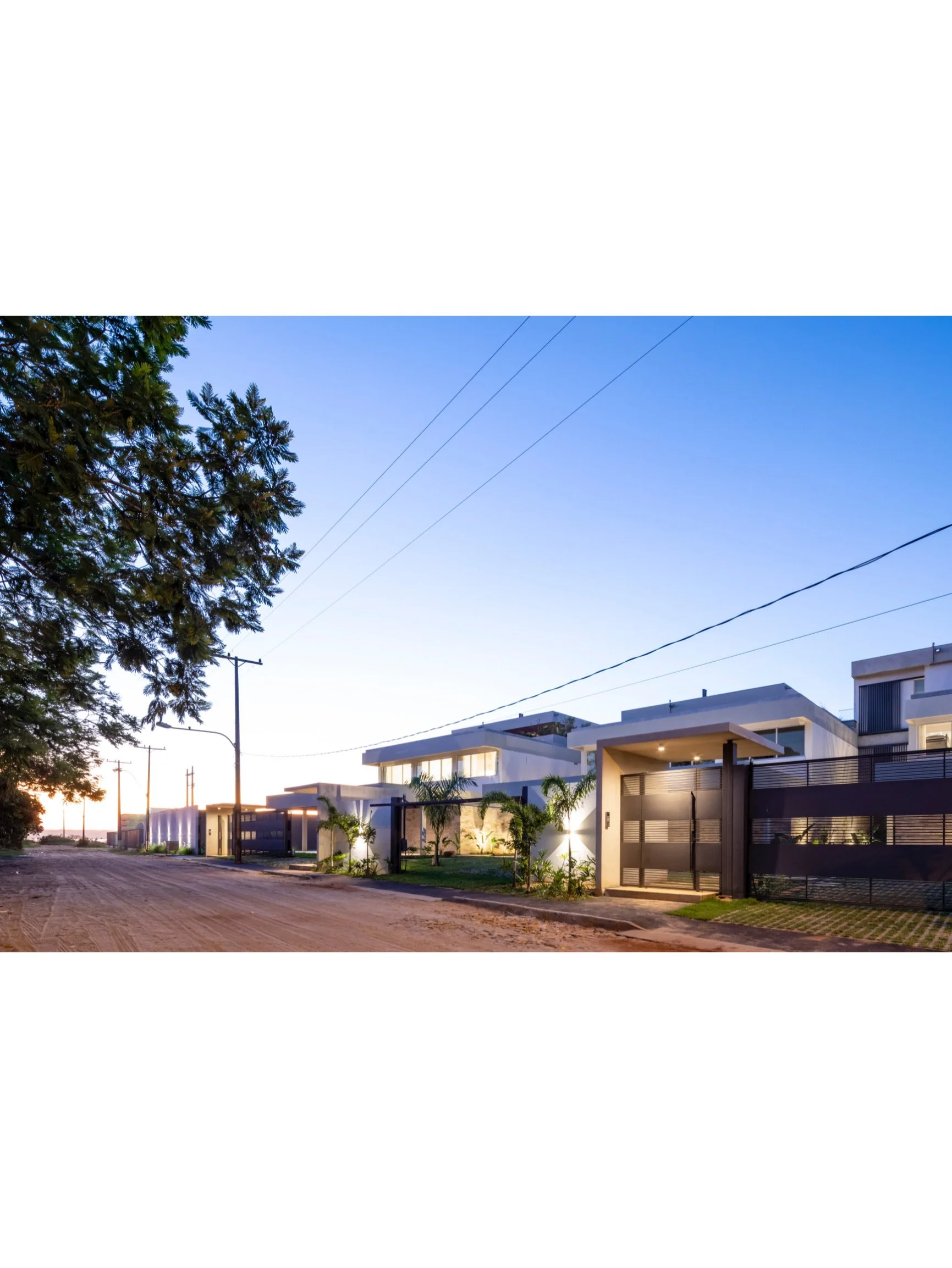 PRO ARQUITECTURA PARAGUAY architecture design PROSA
Luego de la puesta del sol, vista de una calle con casas modernas y jardines con plantas tropicales, en una zona residencial.