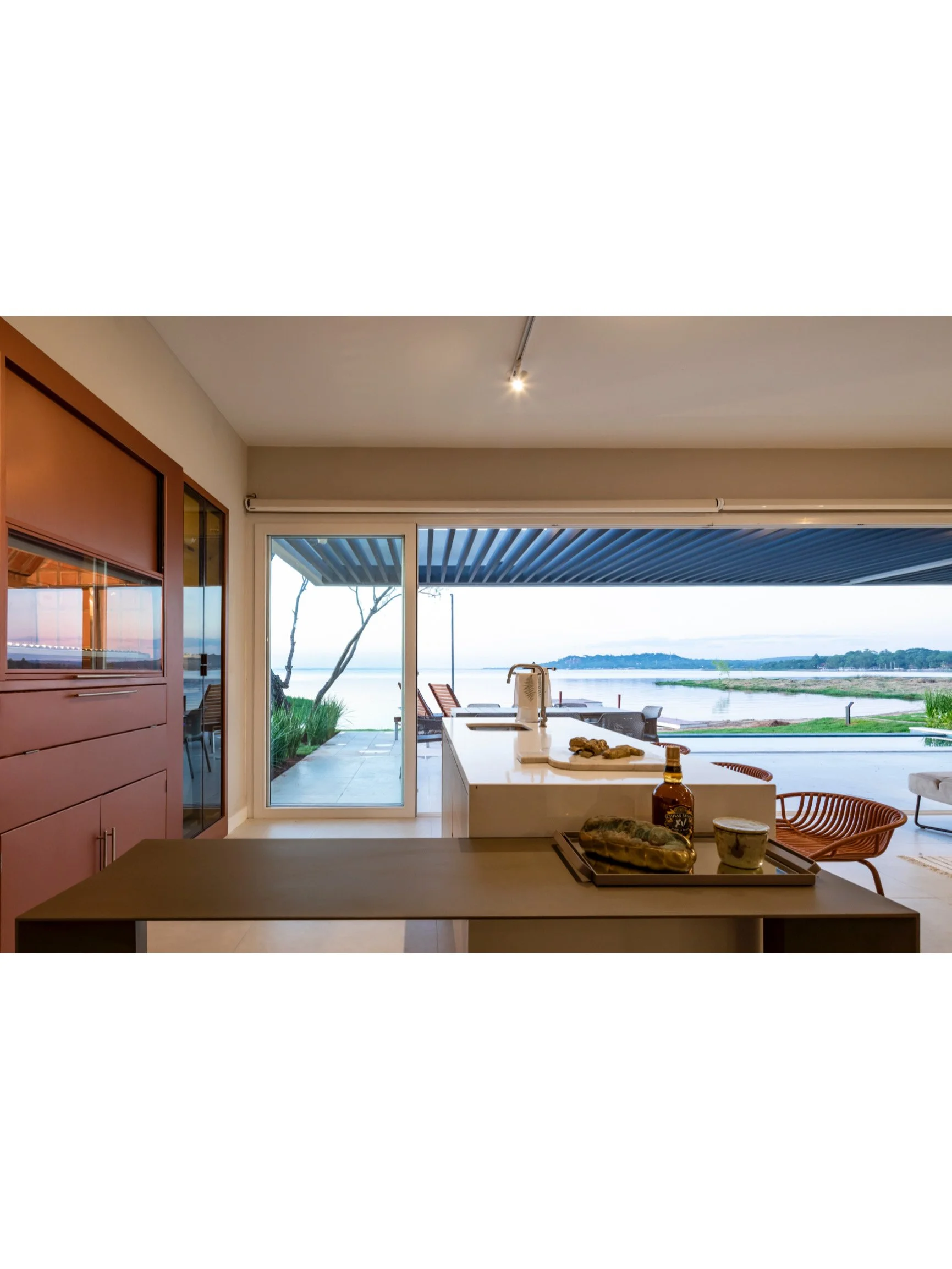 PRO ARQUITECTURA PARAGUAY architecture design PROSA
Vista interior de una cocina moderna con vista al lago, con mesa, sillas de mimbre y comedor al aire libre.