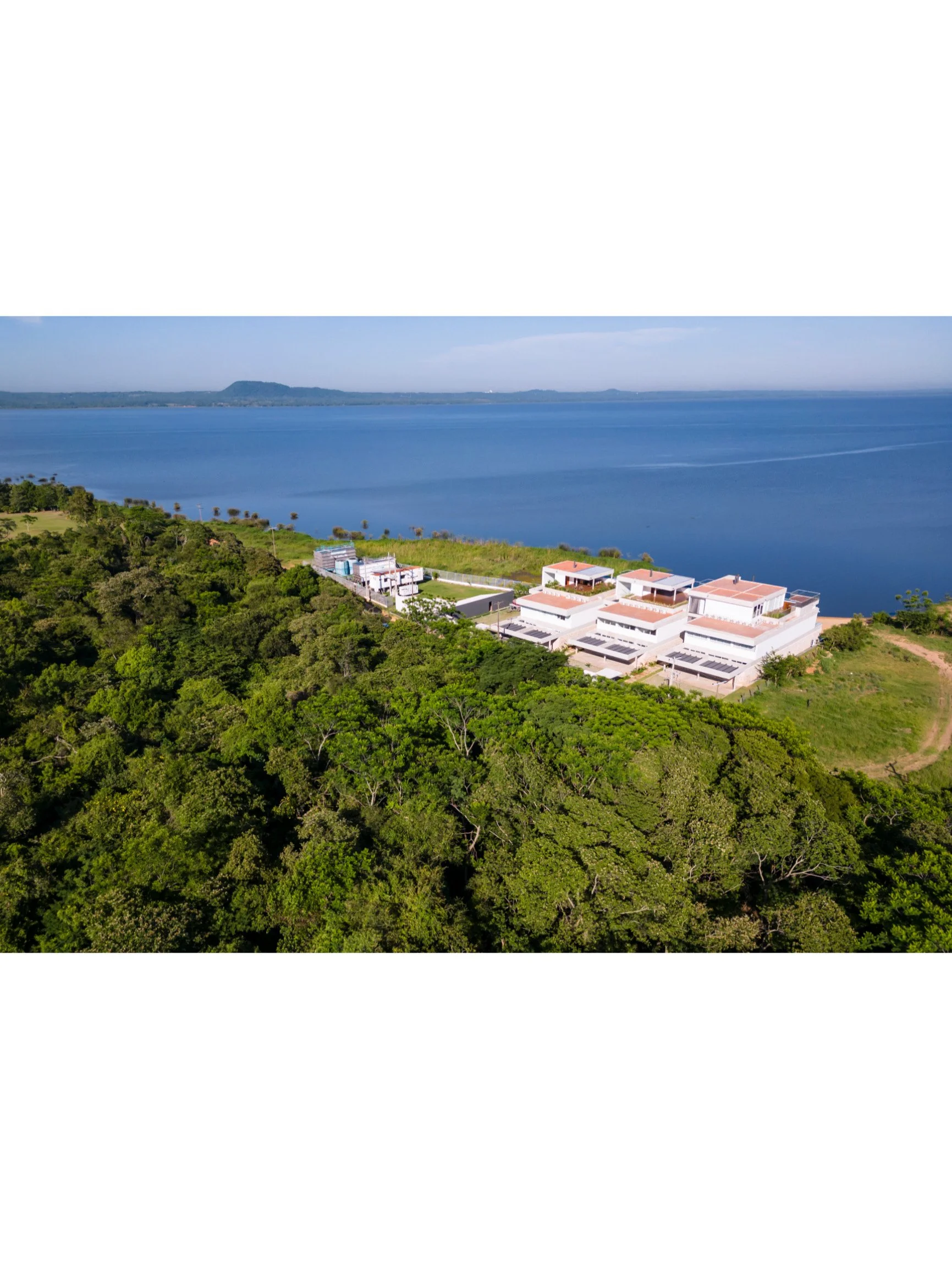 Vista aérea de unos edificios blancos junto a un cuerpo de agua en un entorno natural con árboles verdes y colinas distantes.