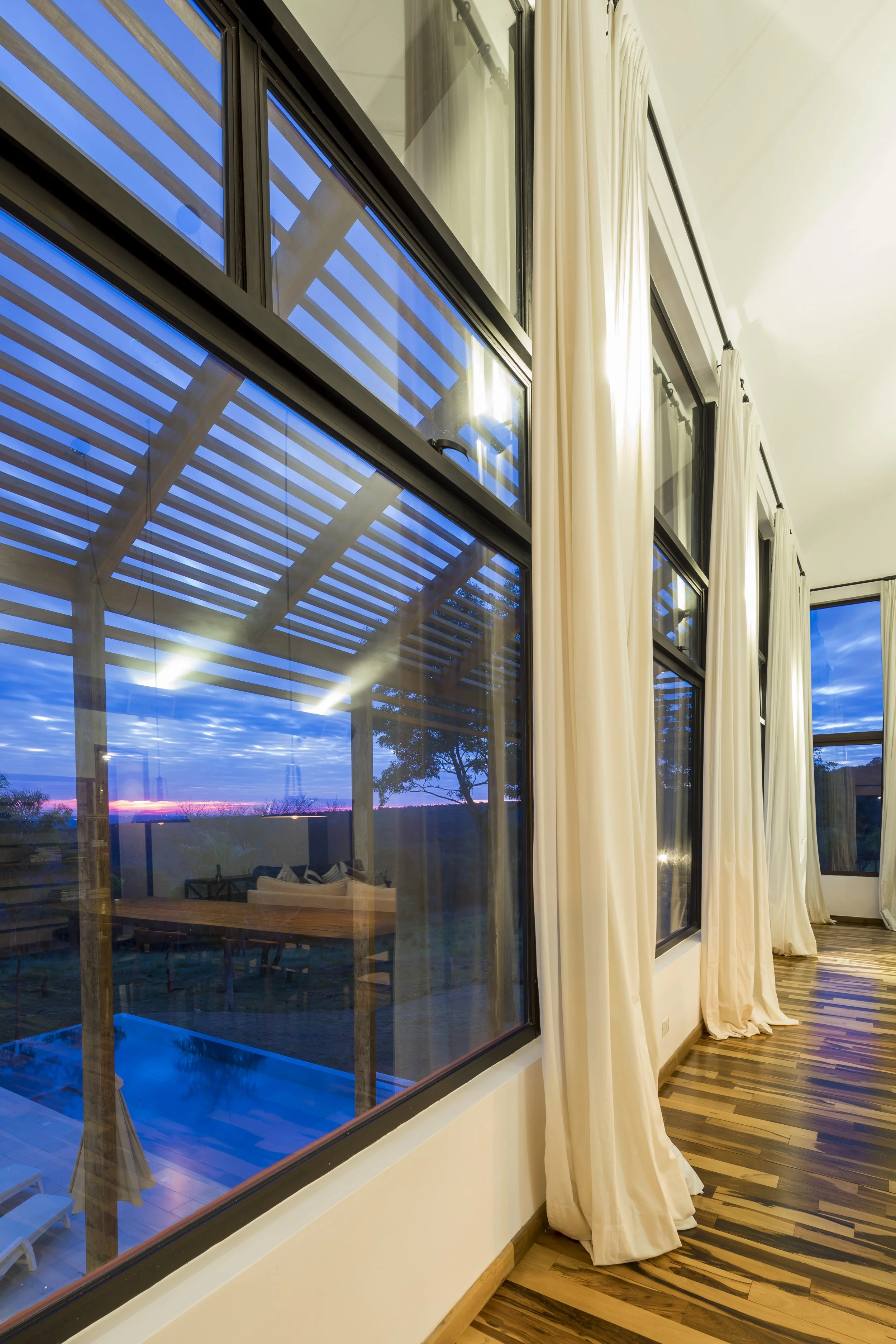 PRO ARQUITECTURA PARAGUAY architecture design PROSA
Vista de una sala con grandes ventanas y cortinas blancas, que muestran un patio exterior con una construcción de madera y un escenario de cielo azul al atardecer.