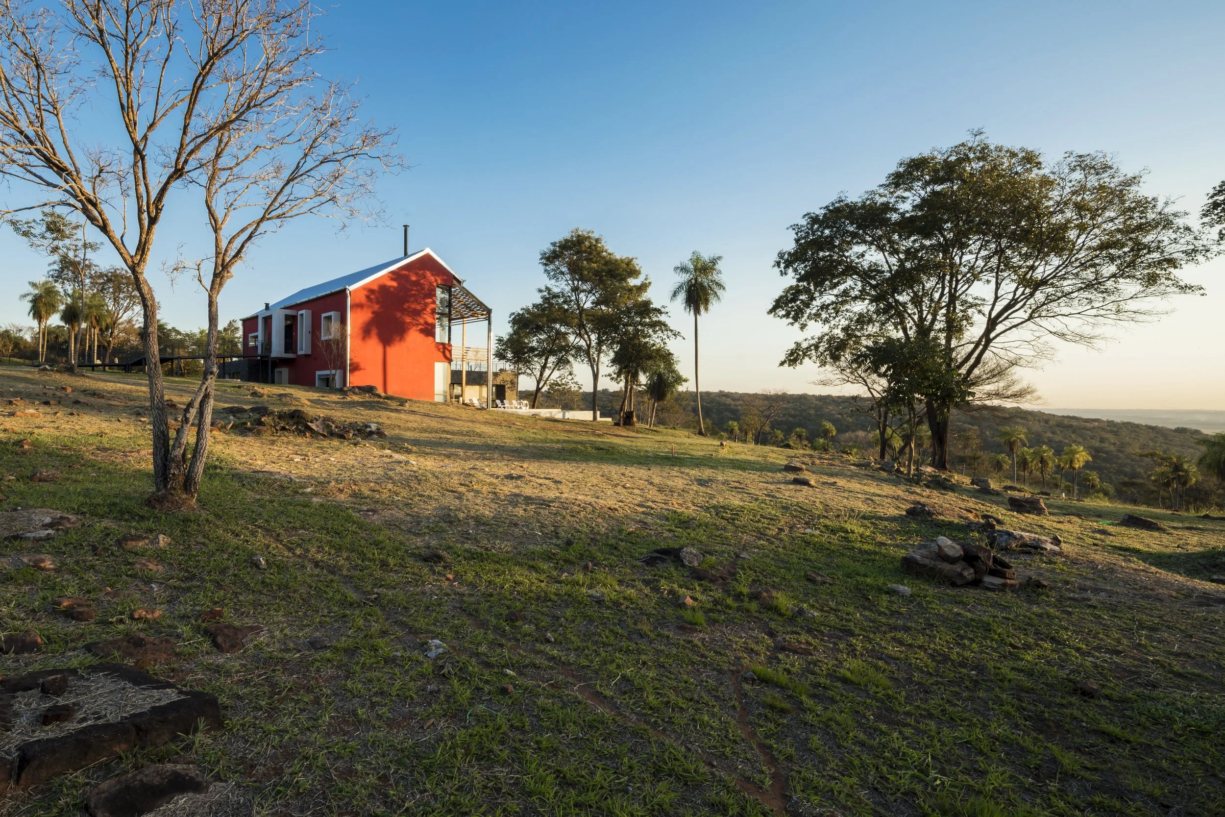 PRO ARQUITECTURA PARAGUAY architecture design PROSA
Casa roja en un paisaje de campo, con árboles dispersos y un cielo despejado al atardecer.