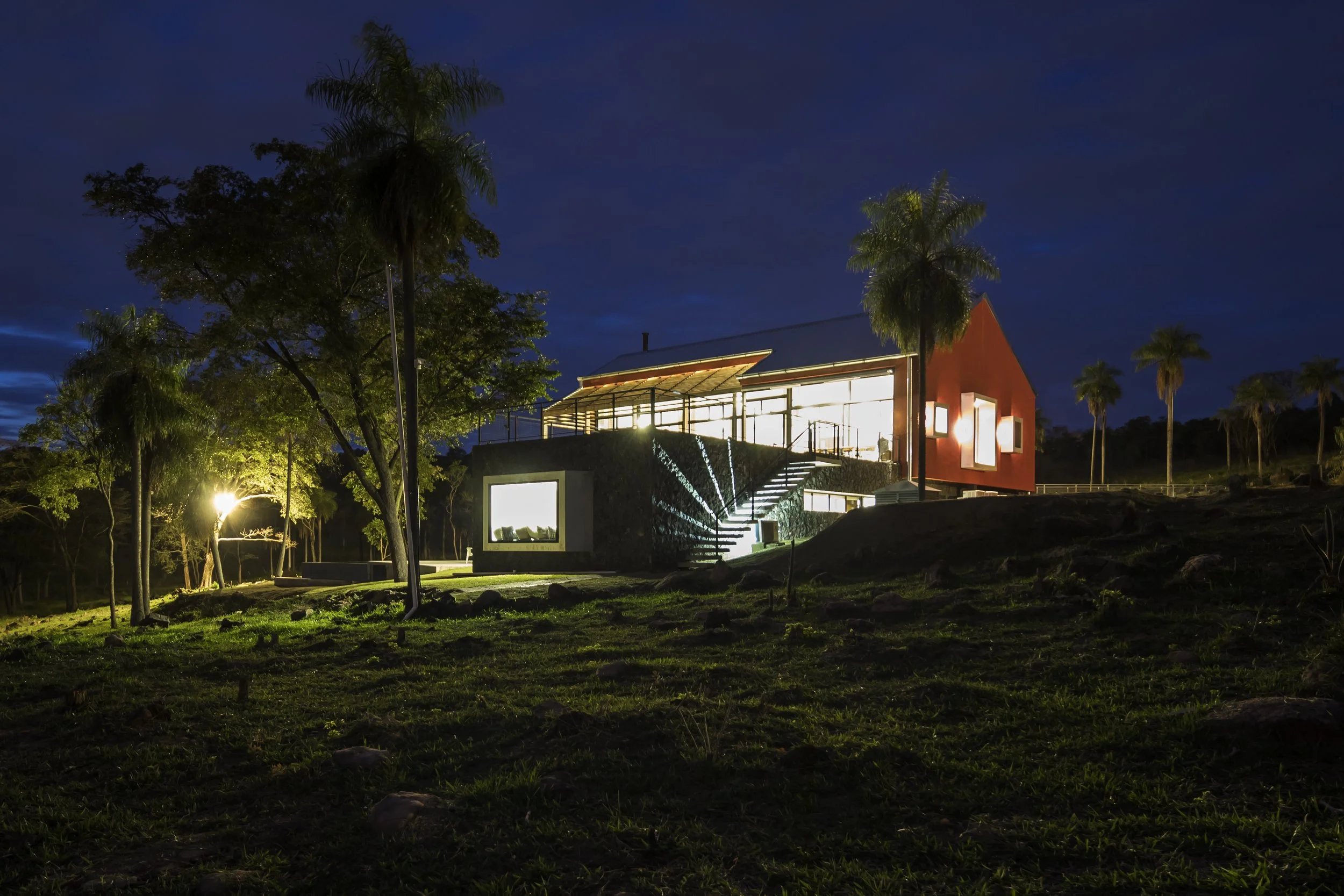 PRO ARQUITECTURA PARAGUAY architecture design PROSA
Casa moderna iluminada en la noche, rodeada de árboles y palmeras, con cielo azul oscuro de fondo.