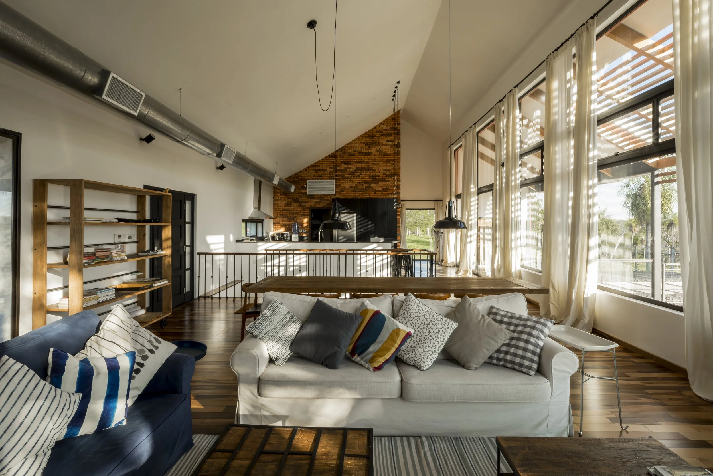 PRO ARQUITECTURA PARAGUAY architecture design PROSA
Sala de estar con sillones, cojines, gran ventanal, cortinas blancas, piso de madera, librero de madera, cocina en el fondo, techo alto con iluminación colgante, pared de ladrillos en el fondo.