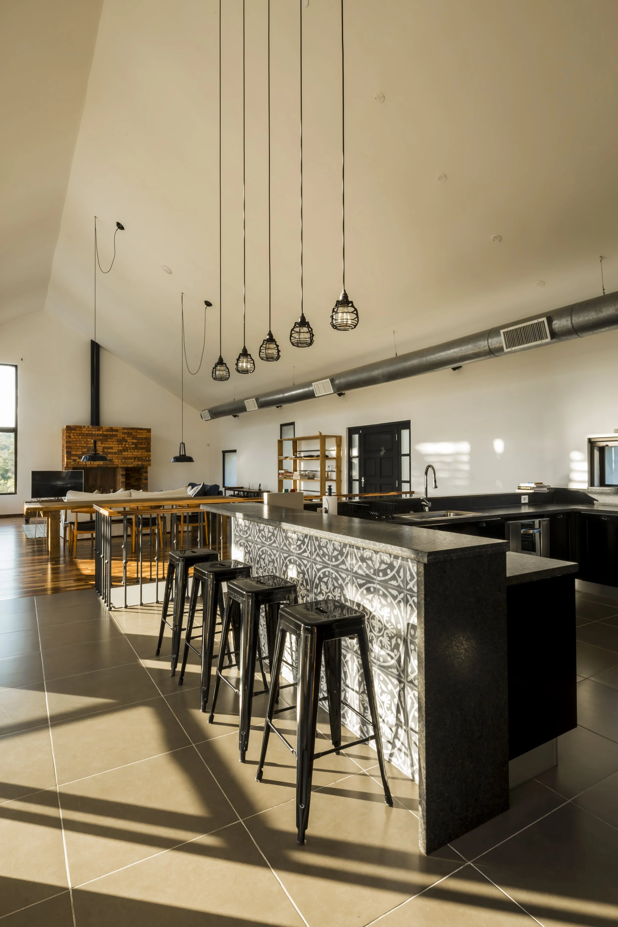 PRO ARQUITECTURA PARAGUAY architecture design PROSA
Cocina moderna con barra de granito, cuatro sillas negras, decoración en tonos neutros, lámparas colgantes y una sala de estar al fondo con chimenea.