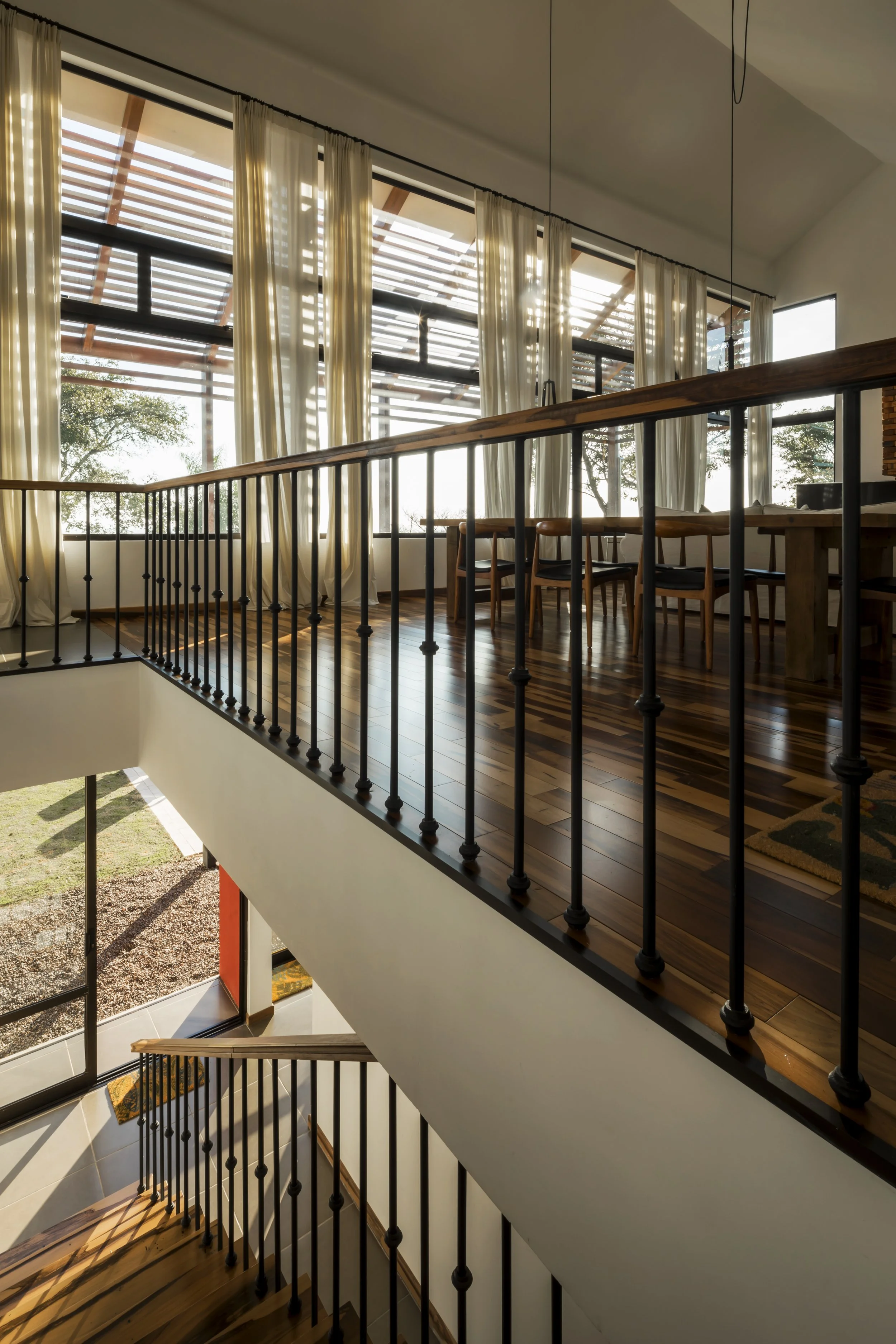 PRO ARQUITECTURA PARAGUAY architecture design PROSA
Vista interior de una casa de dos niveles con barandillas de metal negro, pisos de madera y cortinas blancas en grandes ventanas que dejan entrar luz natural.
