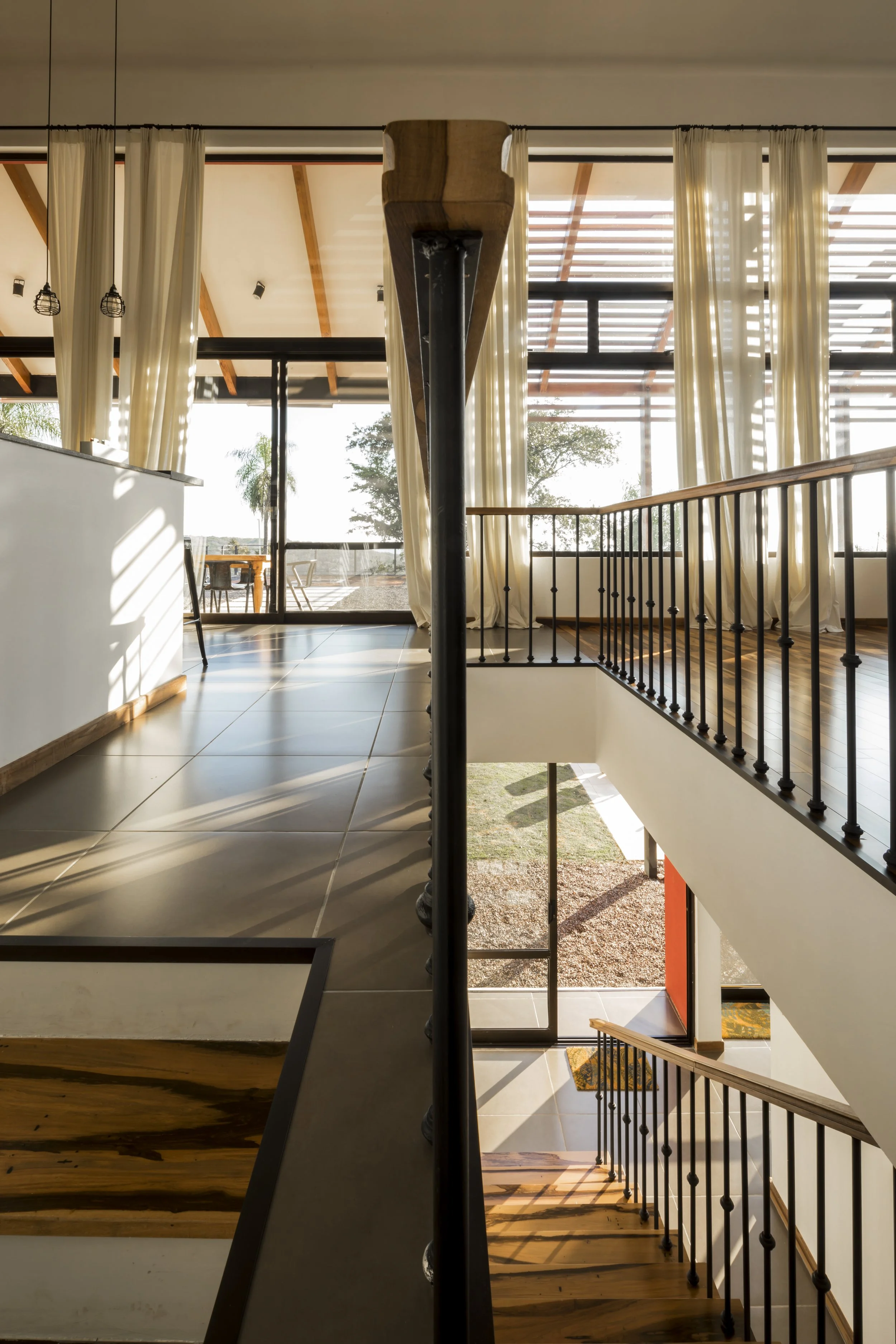 PRO ARQUITECTURA PARAGUAY architecture design PROSA
Vista del interior de una casa moderna con escaleras, barandales de metal, pisos de madera y cerámicas, cortinas blancas y ventanas grandes que dejan pasar luz natural.