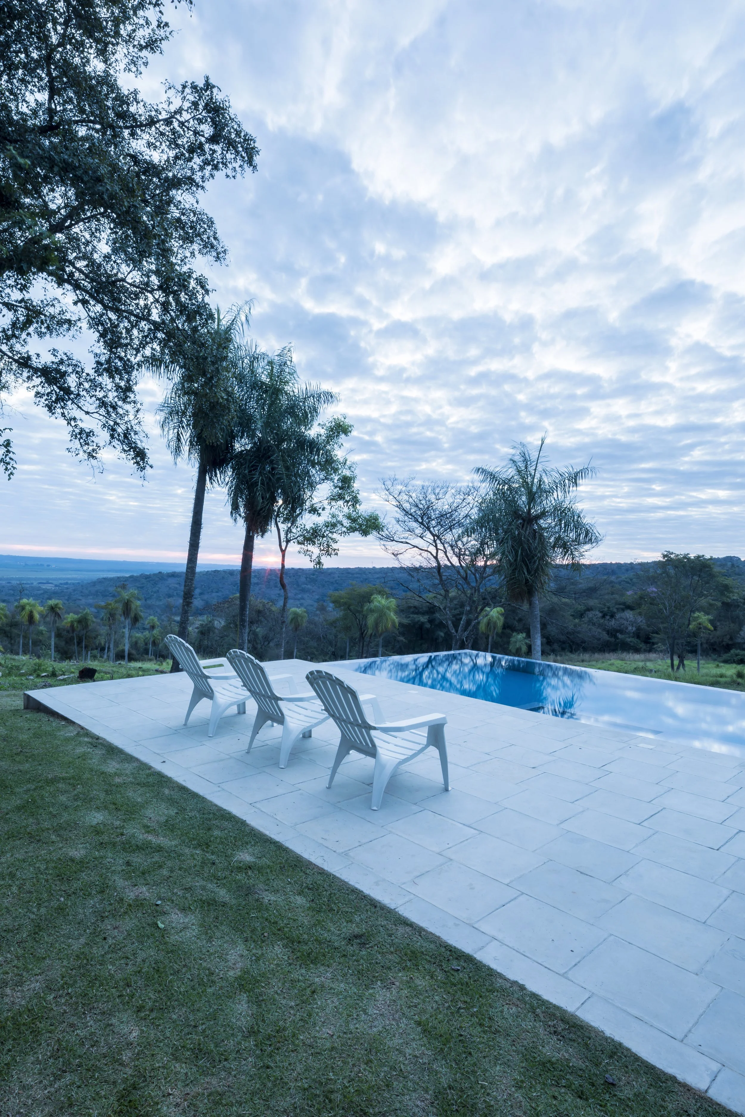 PRO ARQUITECTURA PARAGUAY architecture design PROSA
Piscina en un área de jardín con cuatro sillas blancas, árboles y un paisaje con colinas y cielo nublado al atardecer.